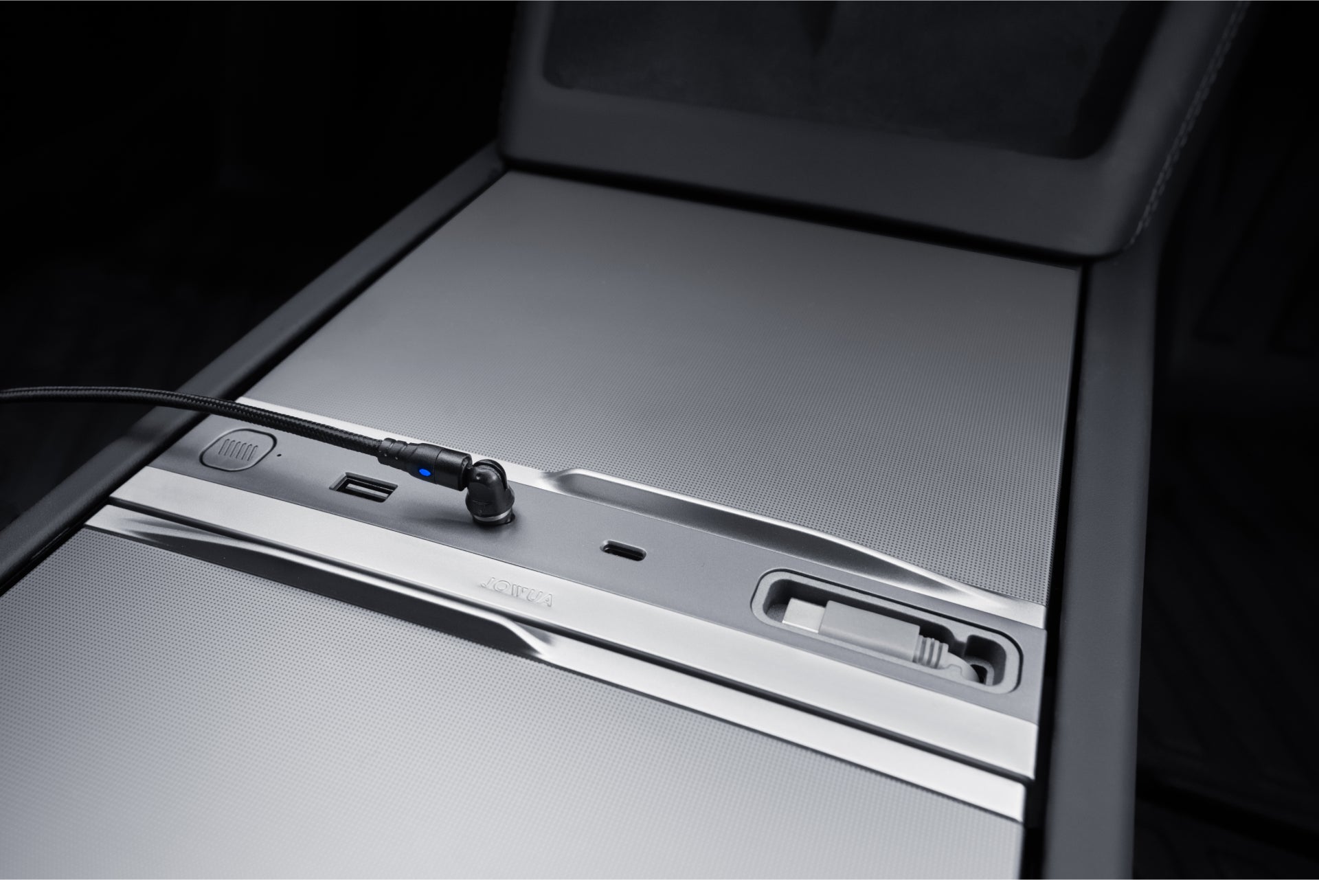 Close-up of a car's center console with a sleek, minimalist design. It features a sliding cover revealing USB ports and a control knob, set against a matte black background.