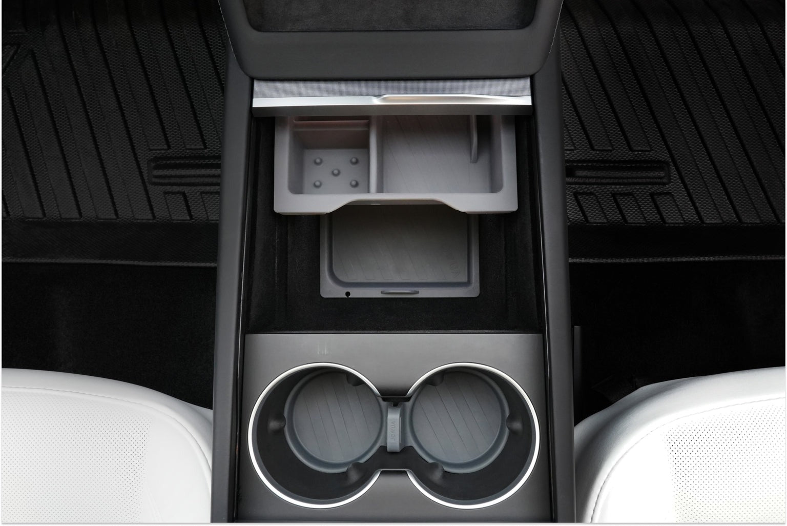 Car interior showing a center console with an open storage compartment and two cup holders, flanked by black and white seats.