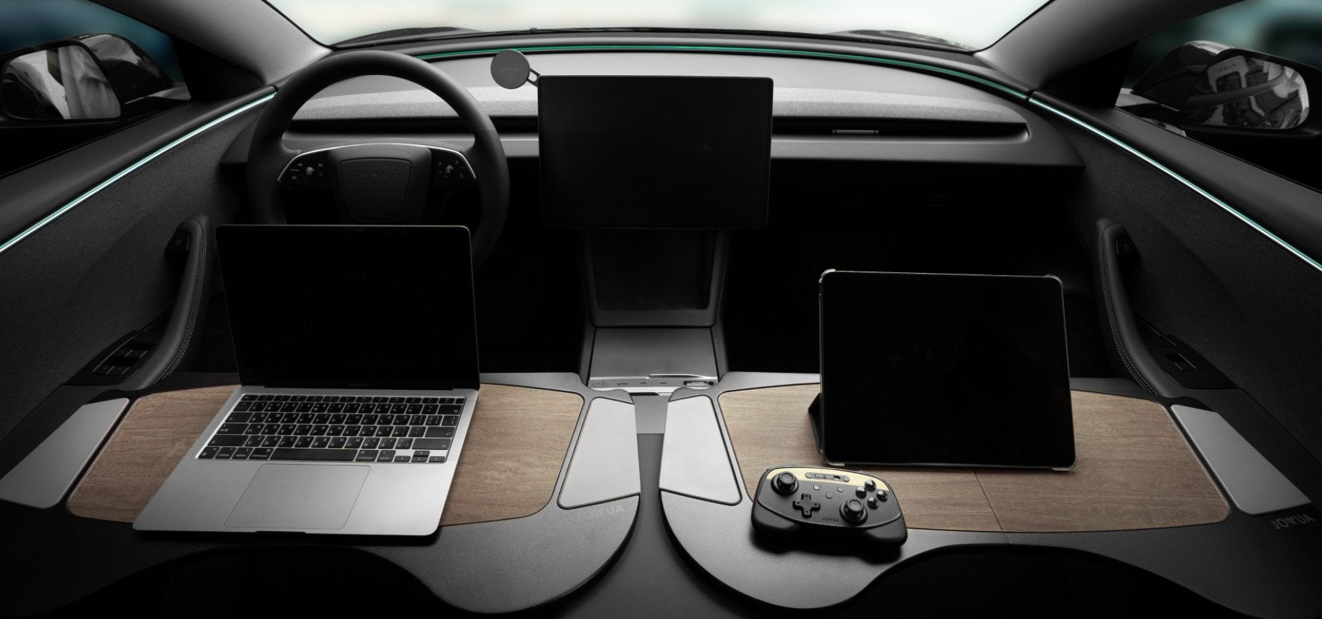 Interior of a futuristic car featuring two seats with wooden trays, each holding electronic devices. A laptop, tablets, and a game controller are visible on elegant black surfaces.