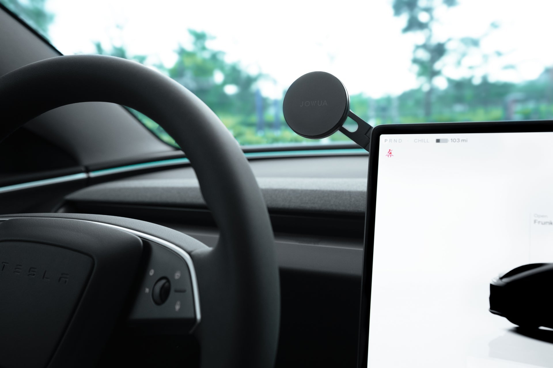 Close-up of a car dashboard showing a round mobile phone mount with a smartphone attached. The car's steering wheel and a part of the digital display are visible, displaying a minimalist screen design.