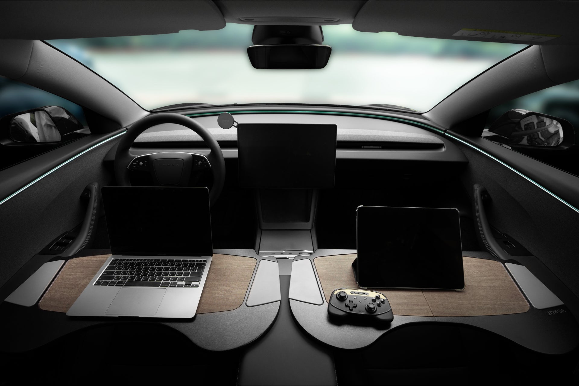 Interior of a car showcasing a futuristic dashboard with two elevated wooden trays holding a laptop and a tablet. A gaming controller rests on the tablet. A central screen is above the console.