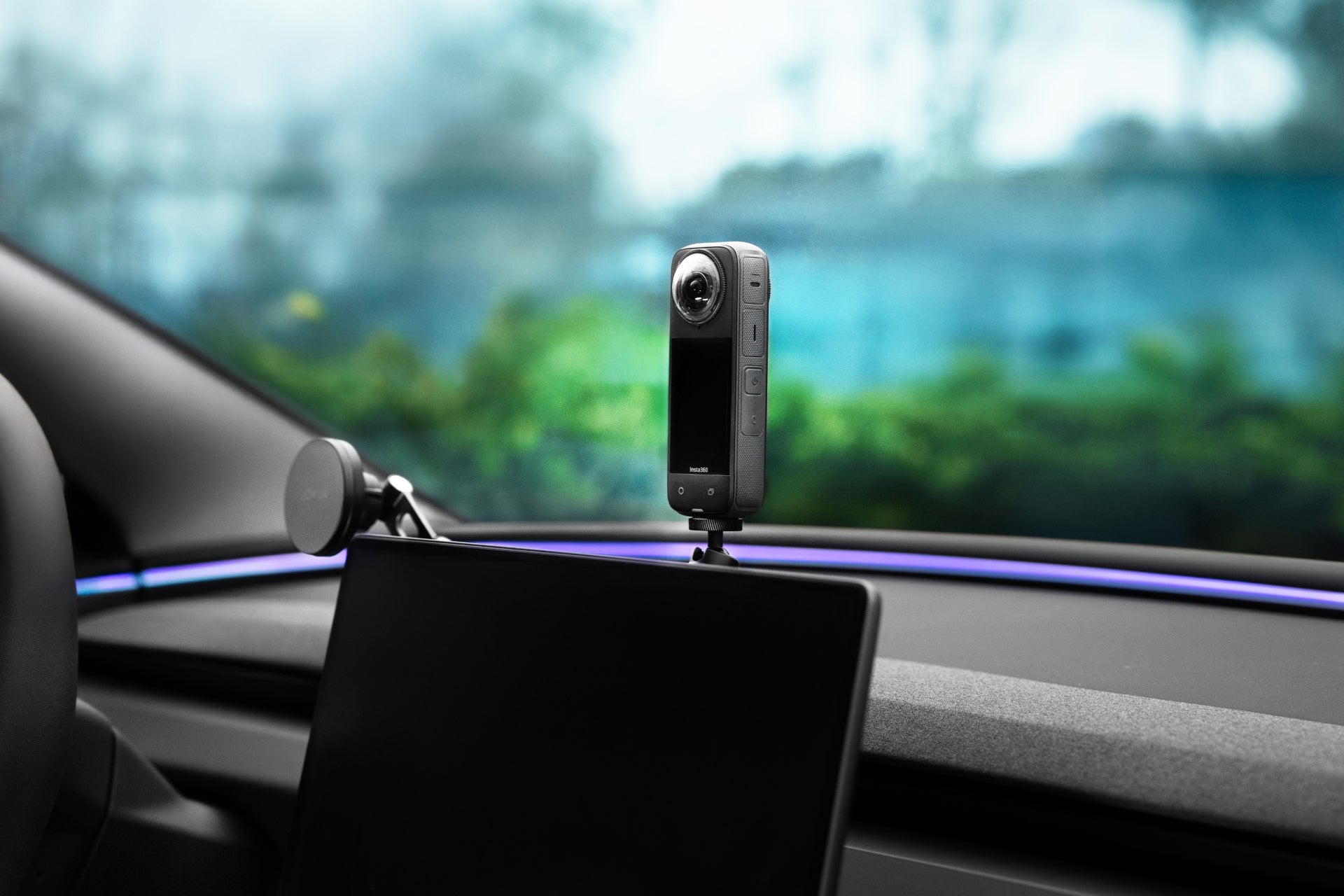 A JOWUA 360° Multi Function Mount, featuring a compact camera with an adjustable ball head, is fixed on a car dashboard. It captures blurred greenery outside the window against a sleek interior with a digital screen—ideal for on-the-go photography.