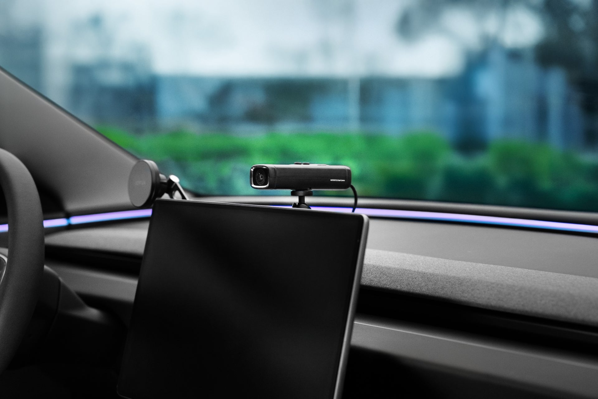 A modern 360° Multifunction Mount by JOWUA, equipped with an adjustable ball head, is placed on the car's dashboard facing the interior. Through the windshield, a blurred view of green foliage and blue sky suggests sleek use for recording video calls or using advanced photography equipment.