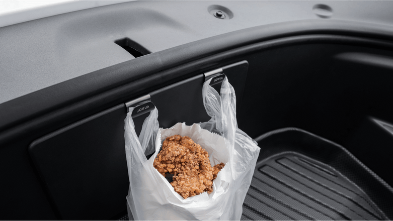 A JOWUA Taiwan Hook Bundle (Model Y) is attached inside a car's storage compartment, holding a plastic bag with a piece of fried chicken. The interior features a dark design and includes a textured rubber mat at the bottom.