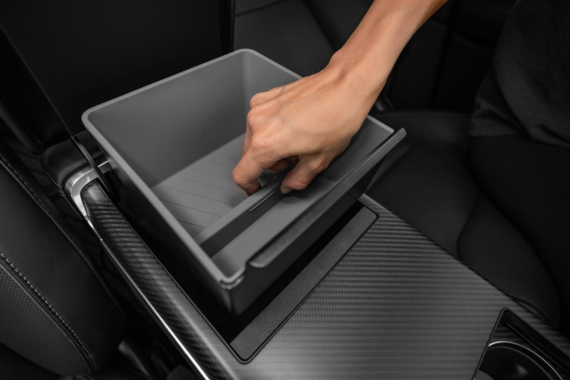 A hand is placing or removing a rectangular, gray JOWUA Armrest Organizer designed for the Tesla Model S/X into a rear compartment of the car's center console. The interior features black upholstery with carbon-fiber accents and premium hybrid materials similar to those found in the Model S/X.