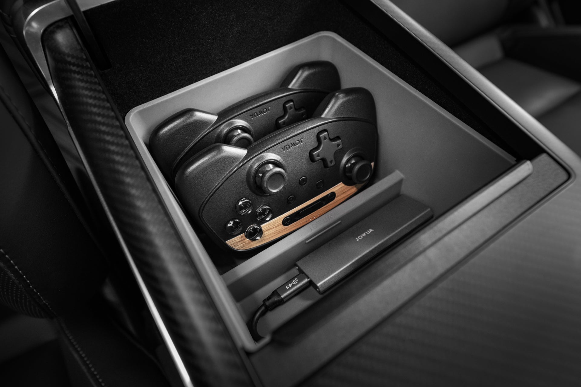 The Armrest Organizer for the Tesla Model S/X, crafted by JOWUA, includes a storage compartment that accommodates two sleek black gaming controllers with prominent joysticks and buttons, along with a wireless charging dock. The interior of this organizer features a premium hybrid material and carbon fiber finish, perfectly aligning with the design ethos of the Model S/X.