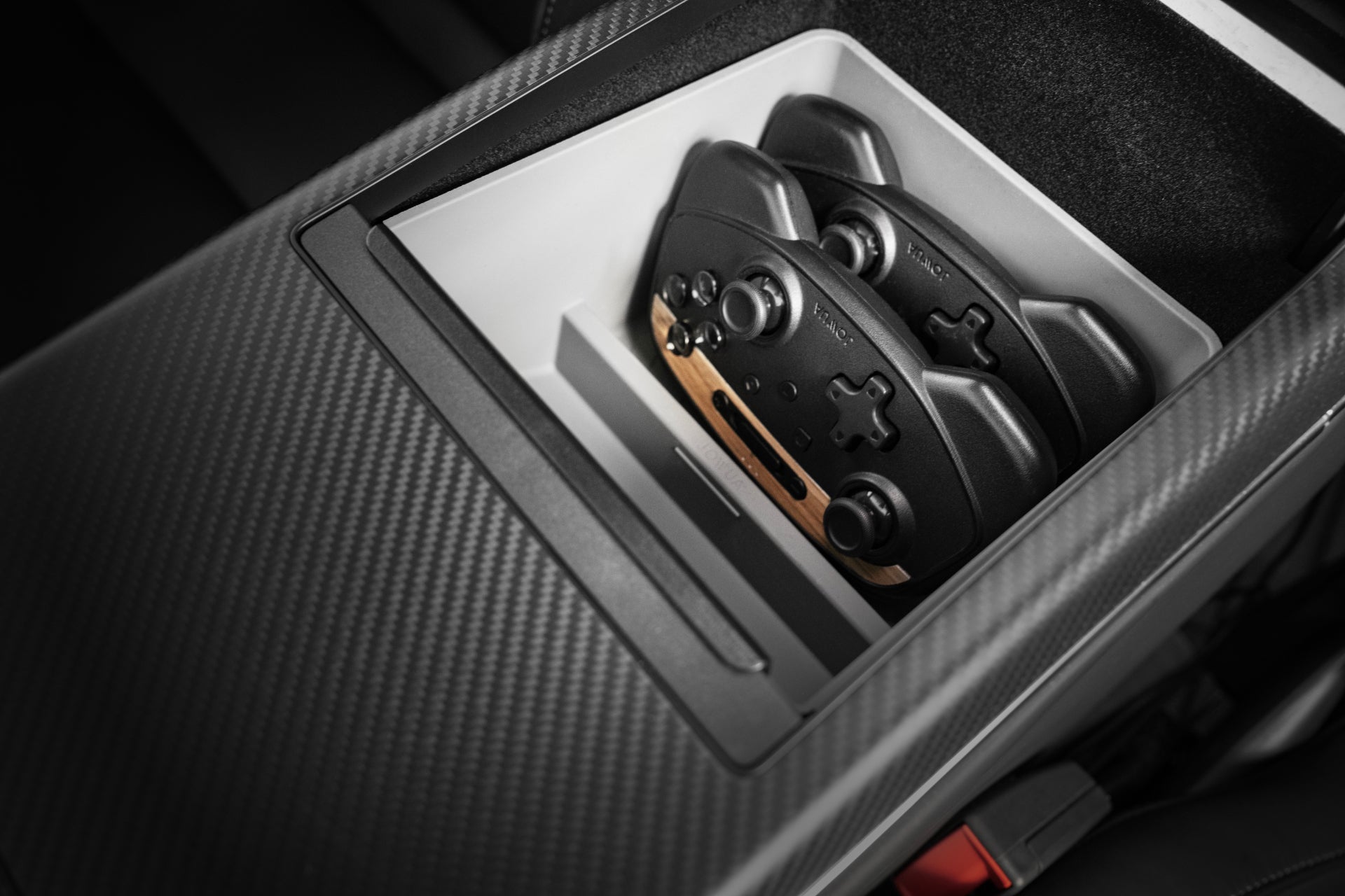 A close-up of the JOWUA Armrest Organizer for Tesla Model S/X showcases two gaming controllers neatly positioned inside. The sleek, gray interior crafted from premium hybrid material highlights a modern car design, complemented by the carbon fiber textured surface surrounding it.