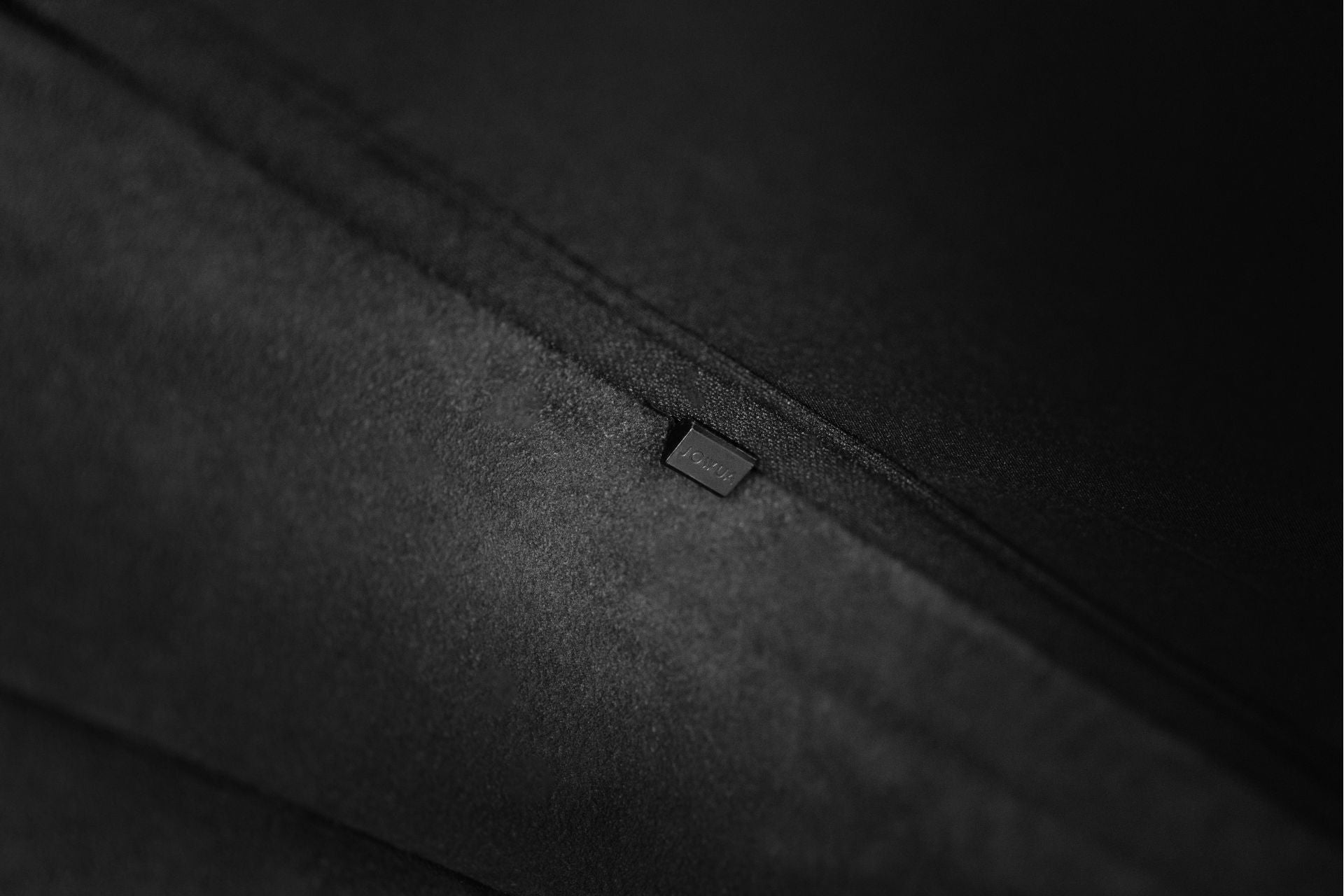 Close-up of the JOWUA Model S Glass Roof Sunshade, featuring a smooth, dark fabric with mesh-like elements and a small rectangular tag stitched along a seam. The texture appears soft and velvety, providing subtle UV protection, while the tag boasts delicate engraving details.