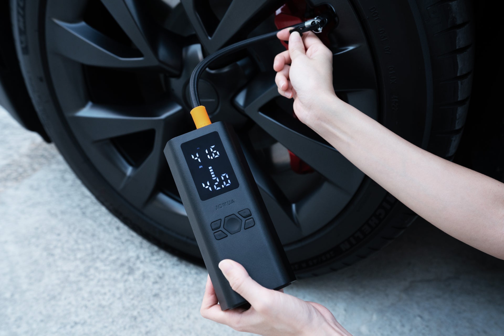 A close-up depicts a person inflating a tire of the JOWUA Model Y Juniper Premium Bundle (Right Hand Drive) using a portable air compressor. The digital display shows numbers as hands skillfully connect it to the valve with the wheel and tread visible in the background.