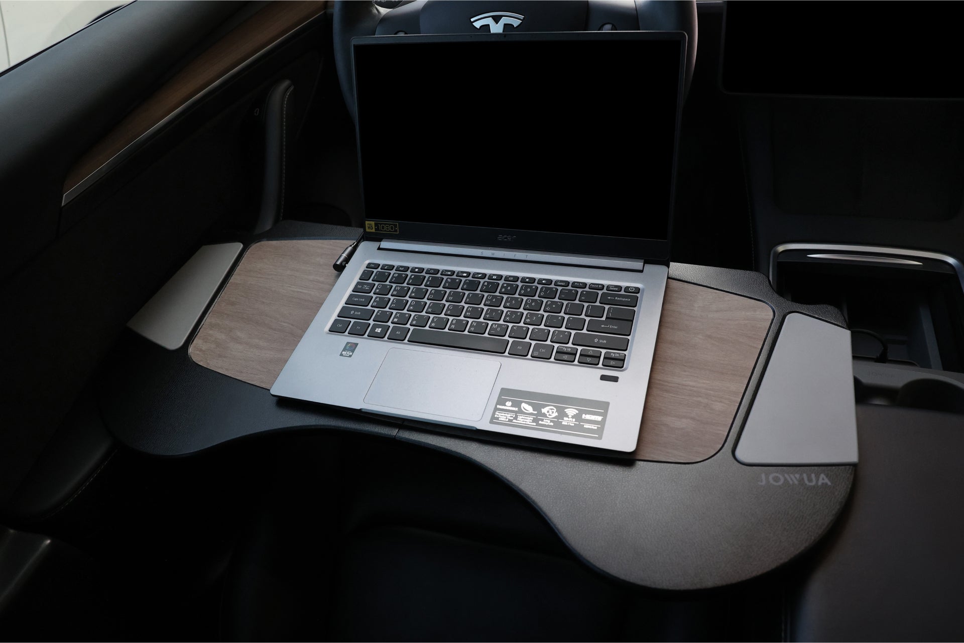 Resting on a custom desk inside the car is a laptop, while JOWUA's 100W USB-C to Magnetic DC Charging Cable ensures stable charging. The desk extends across the front seats, featuring compartments for various items, and its sturdy magnetic design enhances stability. In the background, you can see glimpses of the steering wheel and interior.