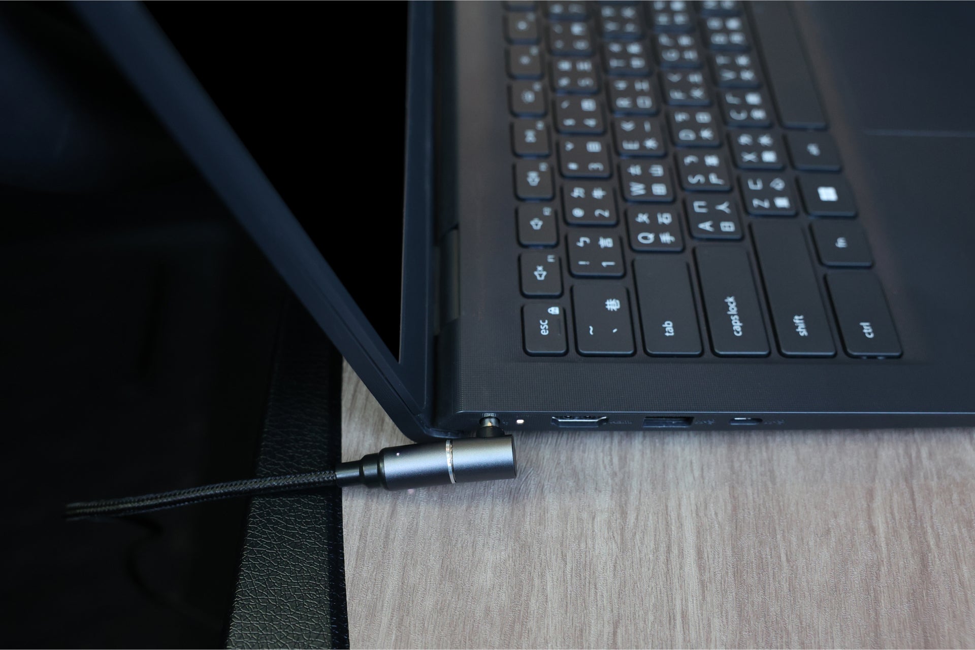 A close-up of a black laptop on a wooden desk highlights the keyboard and a sleek JOWUA 100W USB-C to Magnetic DC Charging Cable connected to the side. The fast charging cable is plugged into one of the laptop's ports, with additional ports visible nearby. The screen is partially open.