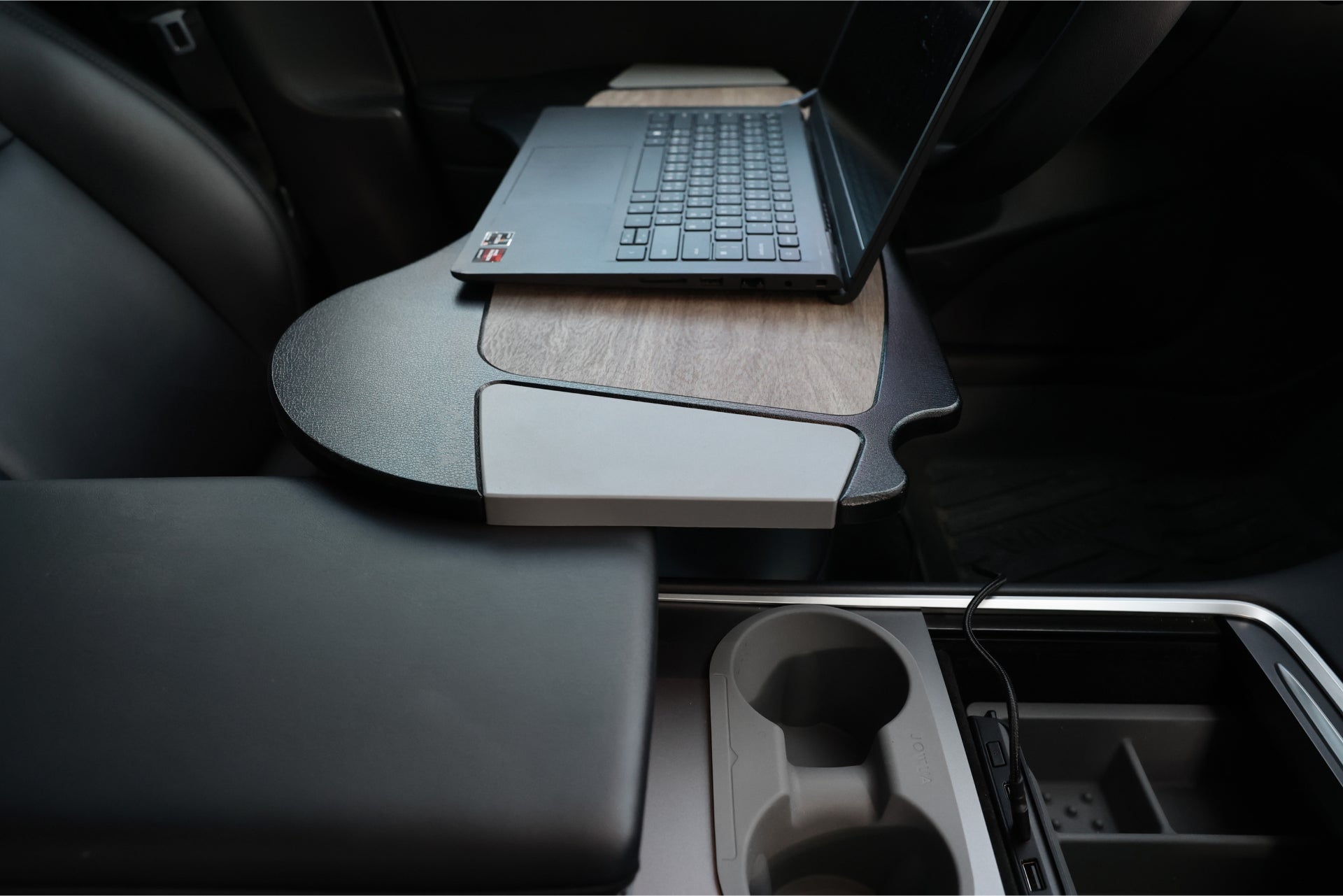 The car interior features a laptop on a custom wooden desk attachment over the front seat, complete with visible cup holders and armrest. The setup includes a JOWUA 100W USB-C to Magnetic DC Charging Cable for fast charging and is crafted from high-quality material for durability.