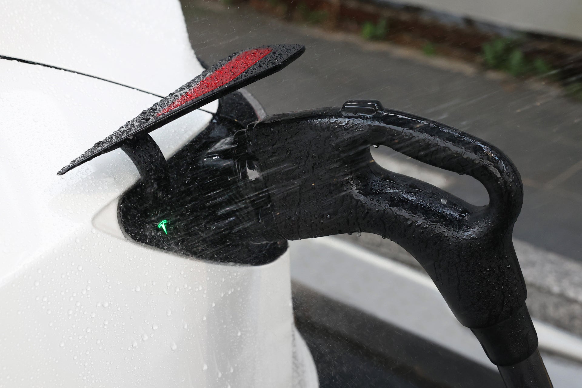 A JOWUA CCS1 to CCS2 Adapter is being used to charge an electric vehicle in the rain. Droplets decorate the sleek black cable and white car, while the charging port remains open and the connector is plugged in securely.