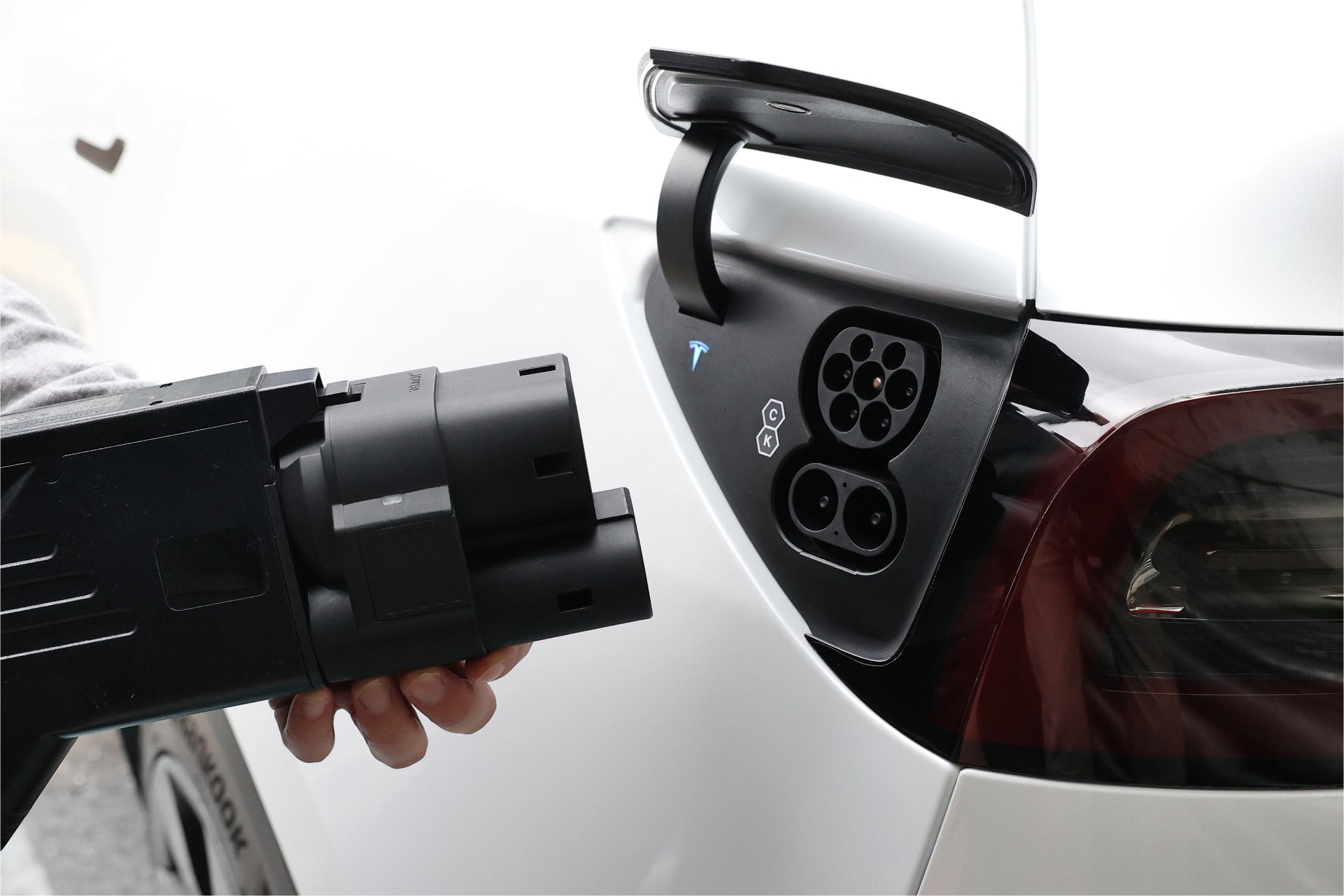 A close-up of a person holding the JOWUA CCS1 to CCS2 Adapter near the charging port of a white car. The port is open, ready for DC Fast Charging. The car's glossy surface reflects light, highlighting its sleek design.