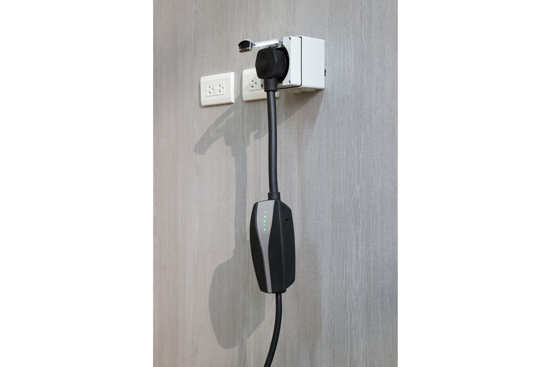 A JOWUA NEMA 14-50 Weatherproof Power Outlet Box is installed against a textured gray wall with a black electric vehicle charging cable plugged into it. The ETL Listed cable extends downward and connects to a charging device with LED indicators, accompanied by three additional power sockets beside the outlet.