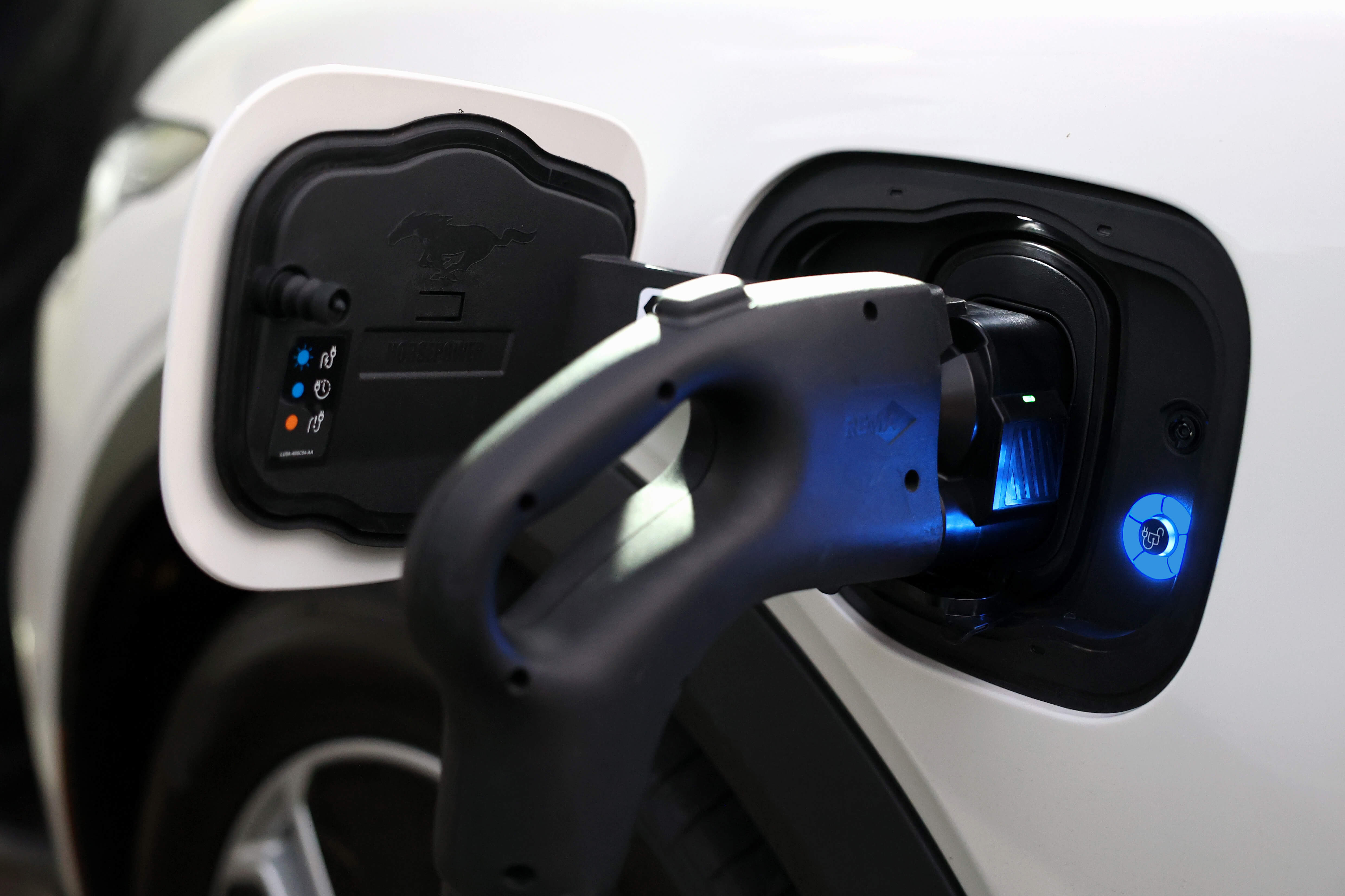 Close-up of a white electric vehicle's charging port with a black charging plug inserted. The charger emits a blue light, indicating it is in operation. A Mustang logo is visible on the charging port cover, and it utilizes a JOWUA CCS1 to CCS2 Adapter for efficient DC Fast Charging compatibility with CCS2 Electric Vehicles.