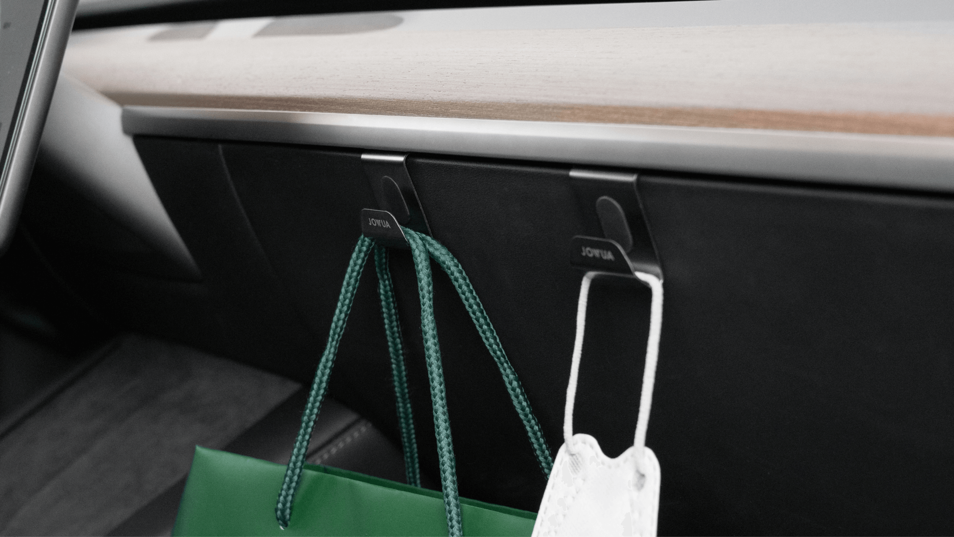 A close-up of a car interior features two JOWUA Taiwan Hook Bundles (Model Y) attached to the dashboard. One hook holds a green shopping bag, while the other secures a white object. The dashboard displays a wooden accent, seamlessly combining functionality with style.