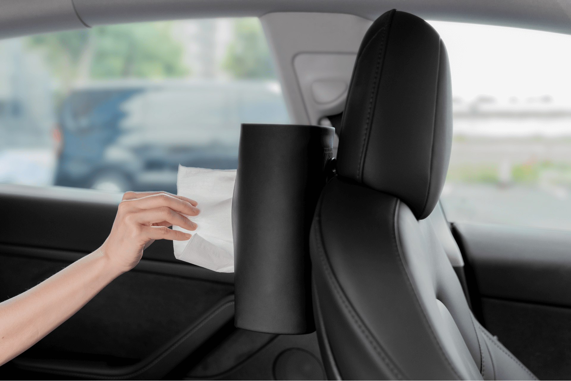 A hand reaches for a tissue from a Hook Bundle by JOWUA Taiwan, elegantly attached to the back of the car's sleek, dark leather seat. Outside, a blurred scene unfolds through the windows, completing the modern and convenient interior.