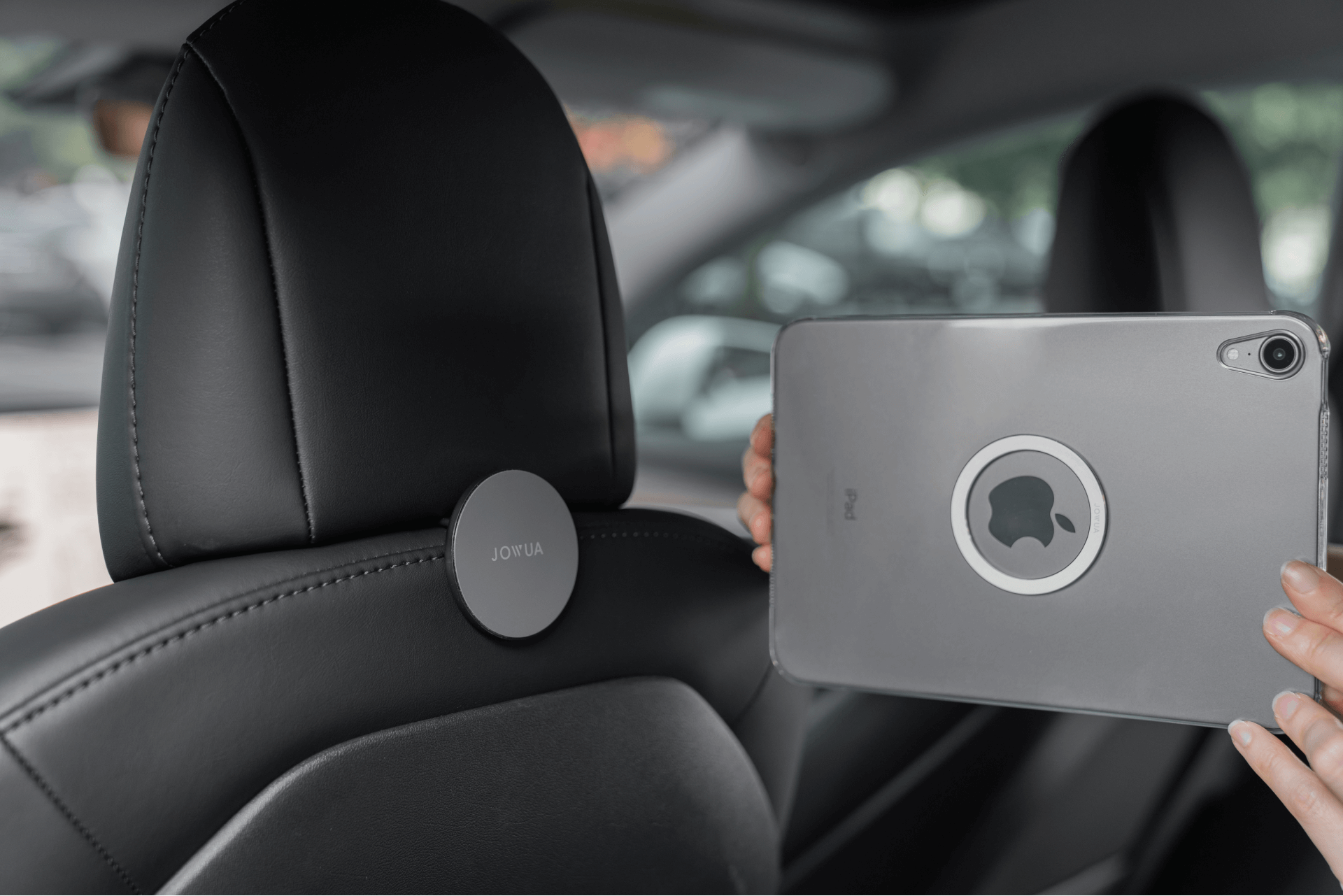 A person holds a tablet with a circular logo on the back, positioned in front of a car headrest where an easy-to-install "Hook Bundle (Model Y)" from JOWUA Taiwan is attached. The interior is sleek and modern, highlighting stainless steel accents, with blurred greenery visible through the window.