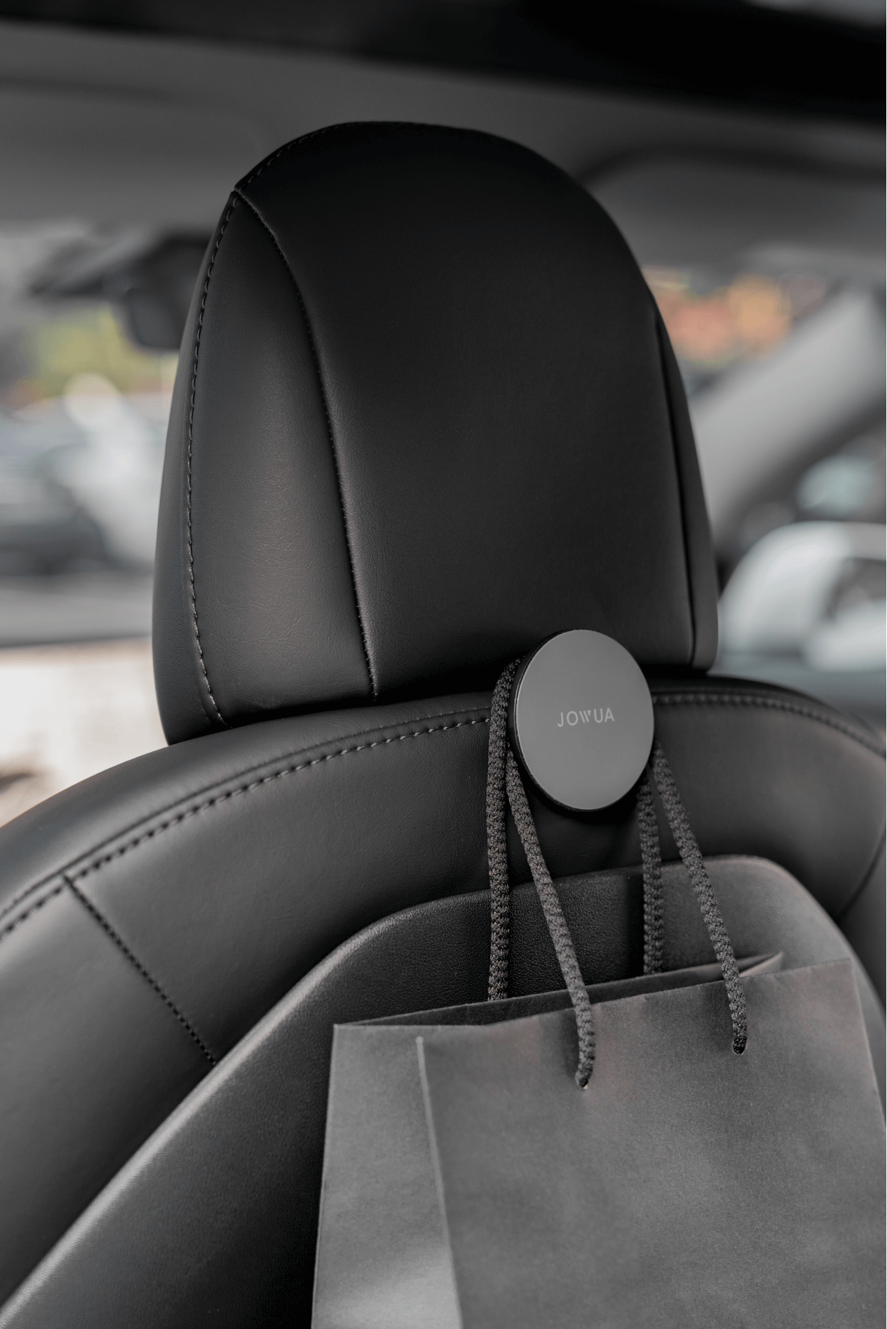 A black car seat is equipped with a compact, round "JOWUA Taiwan" hanger from the Hook Bundle (Model Y), which also serves as a MagSafe car seat holder. A gray shopping bag with black rope handles hangs neatly from it. The background shows a partially blurred view outside the vehicle.