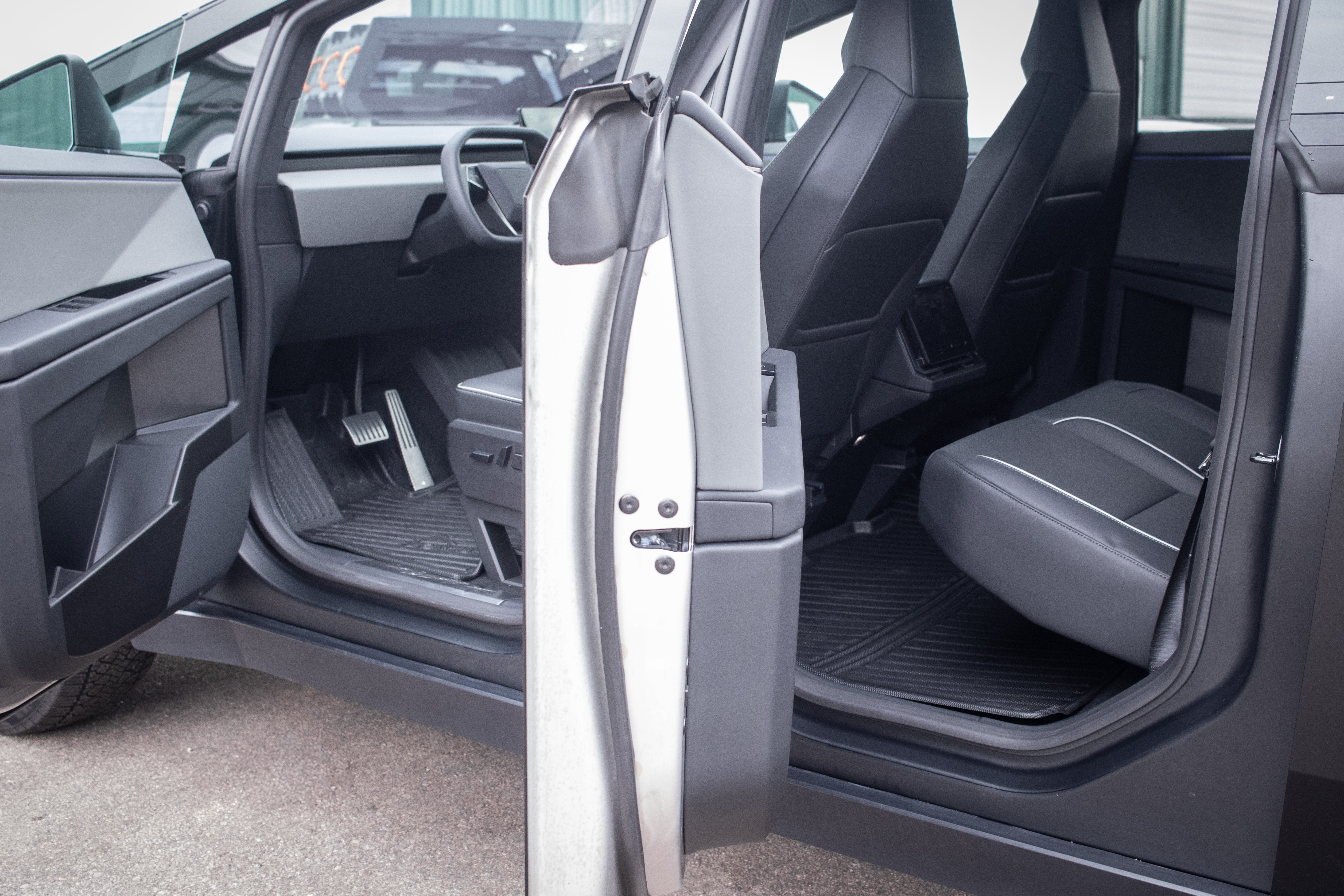 Open the car door to a modern, minimalist interior with dark gray seats, dashboard, and steering wheel. All-weather floor liners from JOWUA add durability, inspired by the Tesla Cybertruck's metallic gray exterior design, as the car rests sleekly on a concrete surface.