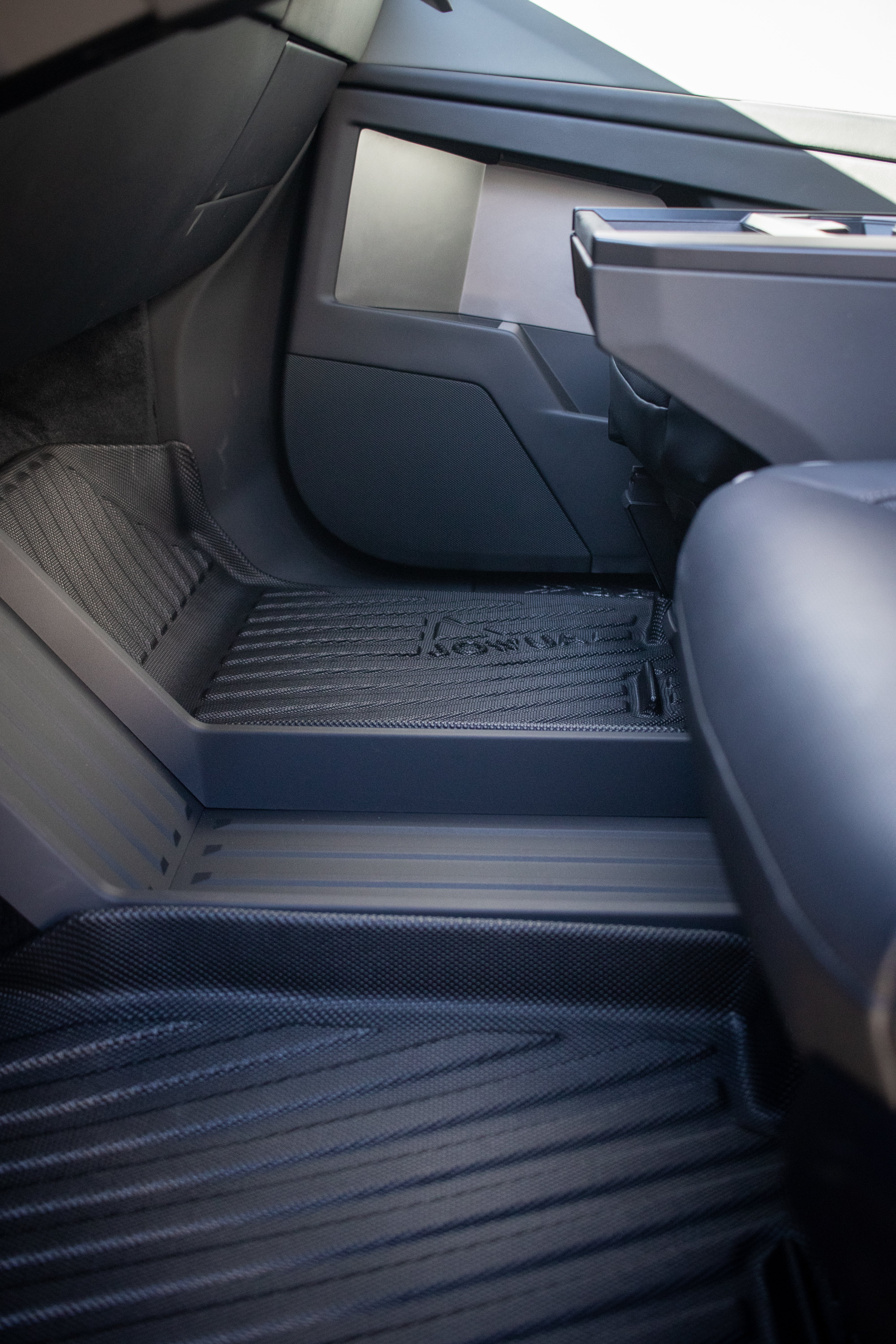 The vehicle's interior features modern, angular designs akin to the Tesla Cybertruck. It focuses on the footwell with JOWUA's All Weather Floor Liners and a sleek, minimalist dashboard for all-weather resilience.