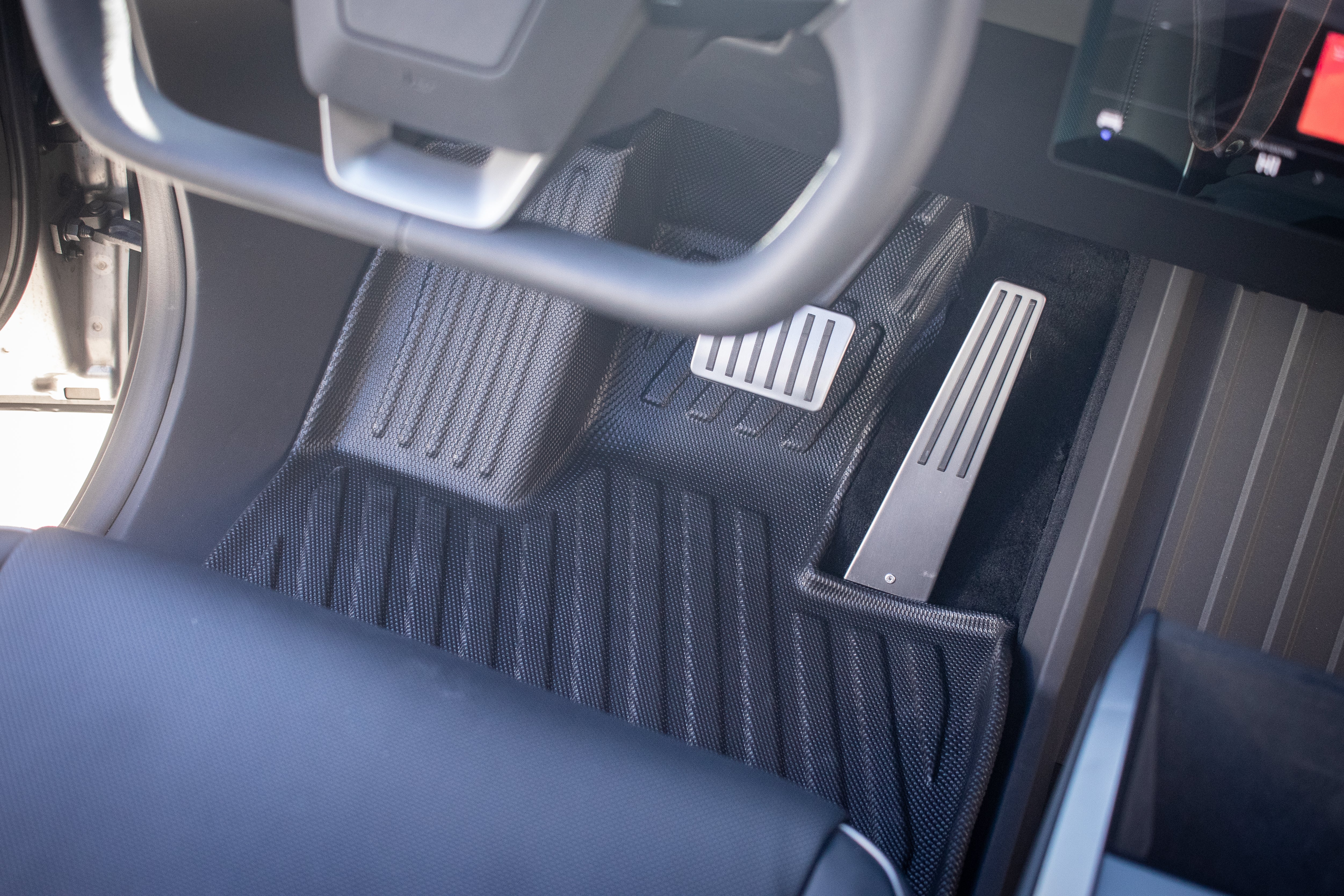 Close-up of a Tesla Cybertruck's driver's area reveals the steering wheel and foot pedals, featuring JOWUA's All Weather Floor Liners with a sleek, modern design. The pedals include both accelerator and brake.