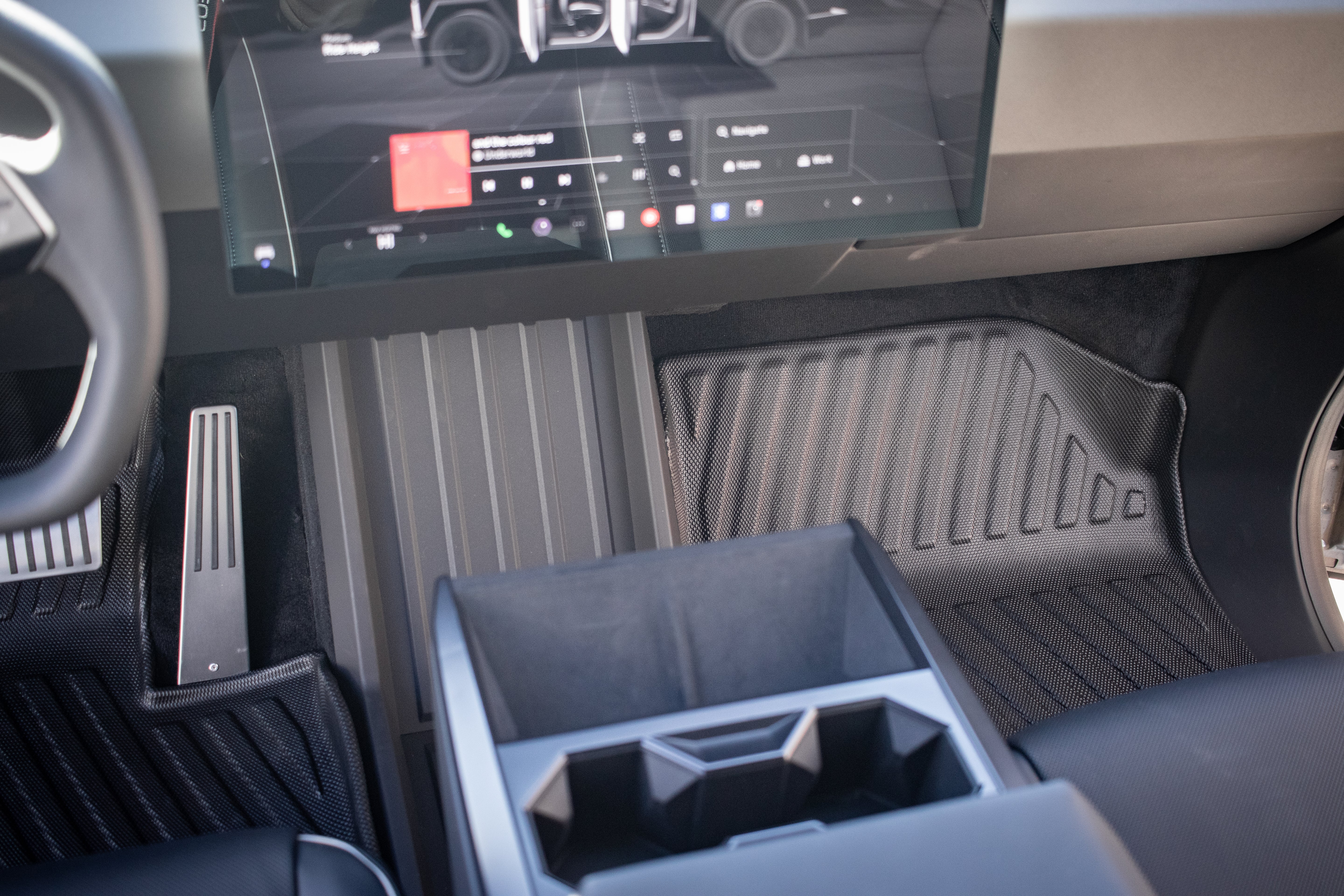 The interior front seat area of the Tesla Cybertruck showcases a large touchscreen with media controls, two pedals, and JOWUA's All Weather Floor Liners. The center console includes cup holders, a storage compartment, and an all-weather liner for durability.