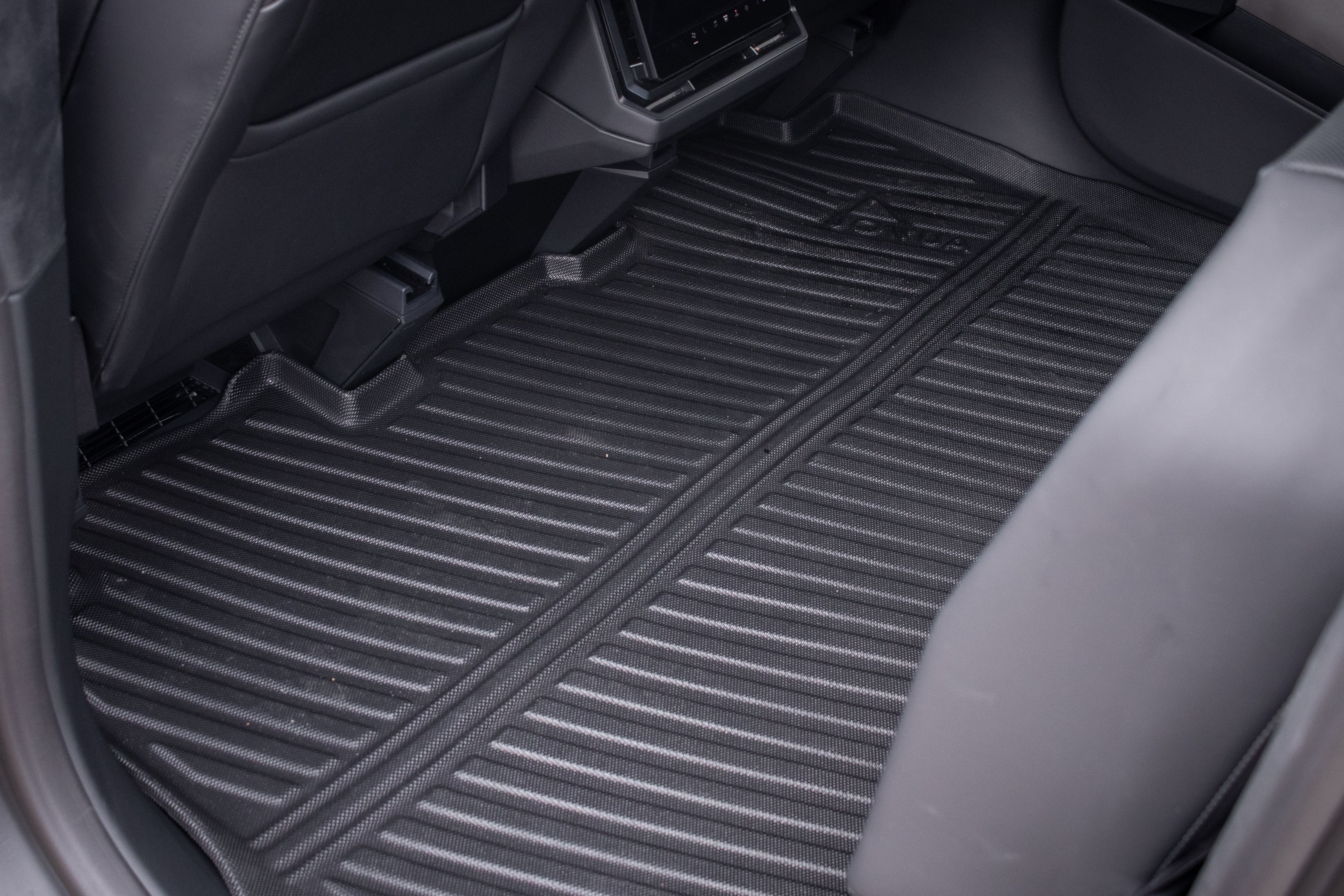 The image displays the rear footwell of a car equipped with JOWUA black rubber all-weather floor liners for the Tesla Cybertruck, featuring grooves and the brand imprint. The car seats and interior, adorned in dark upholstery, enhance the sleek design aesthetic.