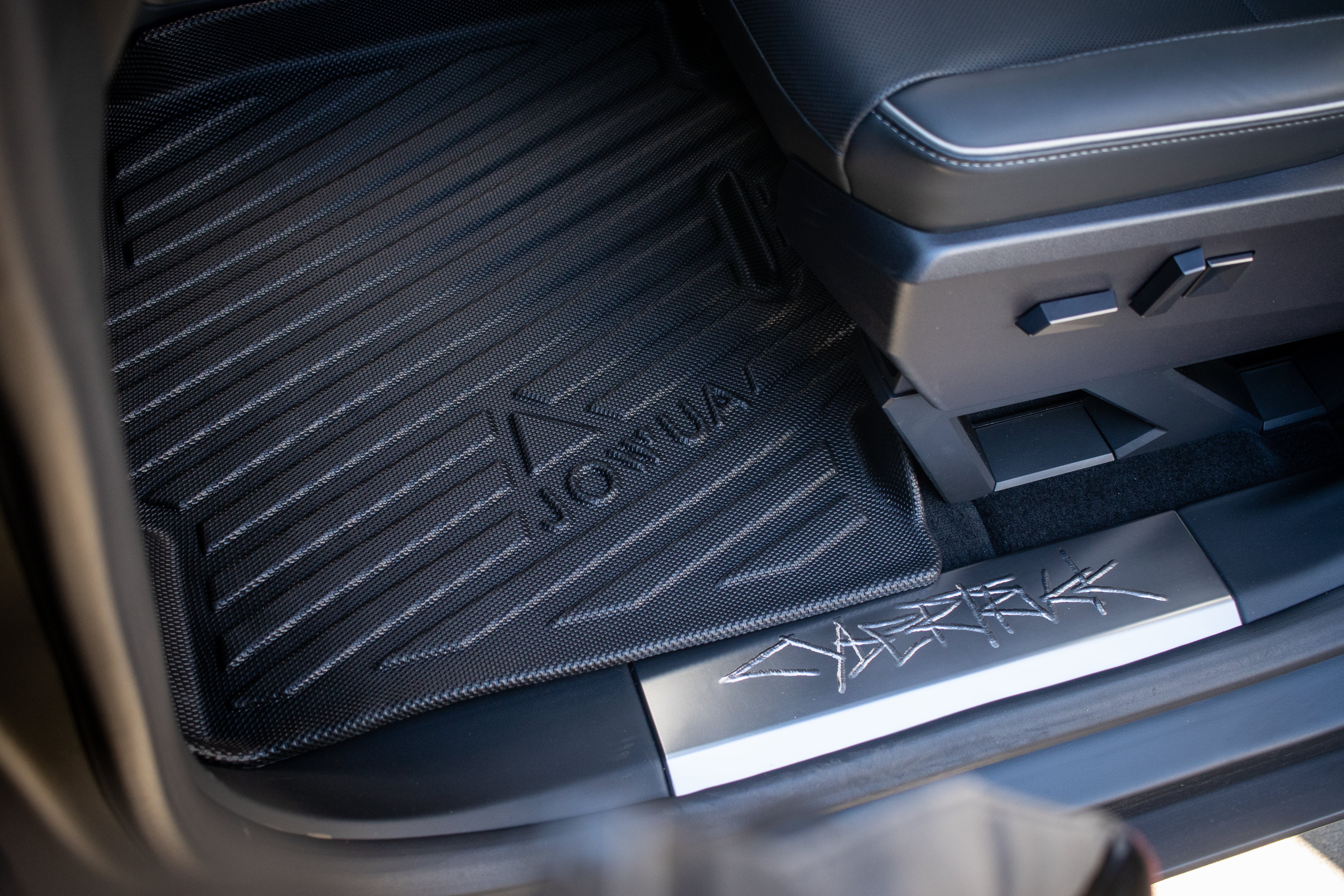 Close-up of a car's interior featuring JOWUA All Weather Floor Liners (Tesla Cybertruck) with a textured, ridged pattern. The liner bears the brand name "JOWUA". Nearby, the car's door sill displays a design inspired by the rugged Tesla Cybertruck aesthetic.