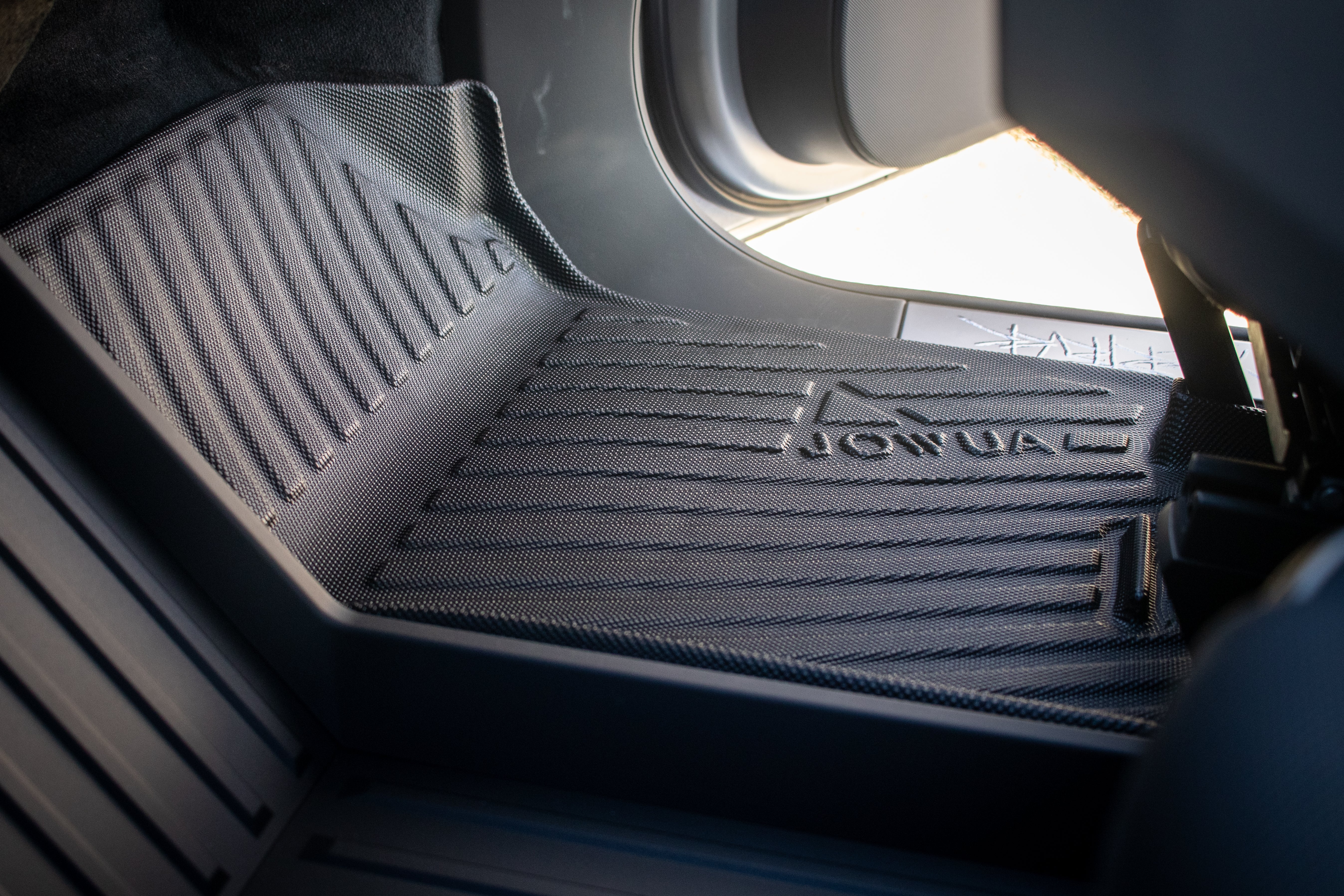 A close-up of a black textured JOWUA floor mat, labeled "All Weather Floor Liners (Tesla Cybertruck)," sits in the footwell with raised sides and a ribbed pattern trapping dirt. Sunlight highlights part of it through the open car door.