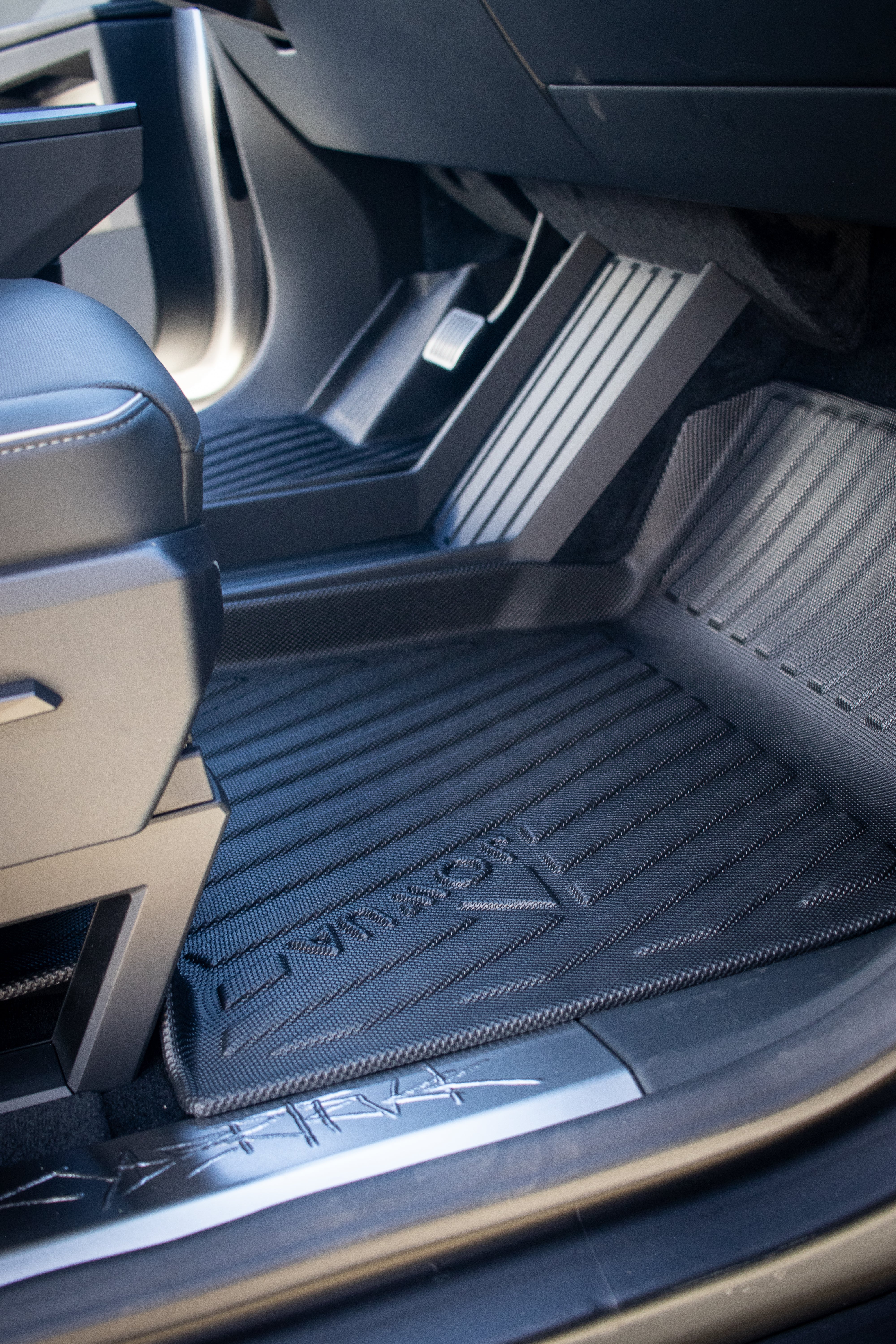 A close-up of a car interior features JOWUA's sleek and modern All Weather Floor Liners for the Tesla Cybertruck, showcasing black, ribbed design with an embossed logo. The driver's seat and pedals are partially visible, highlighting durability and style.