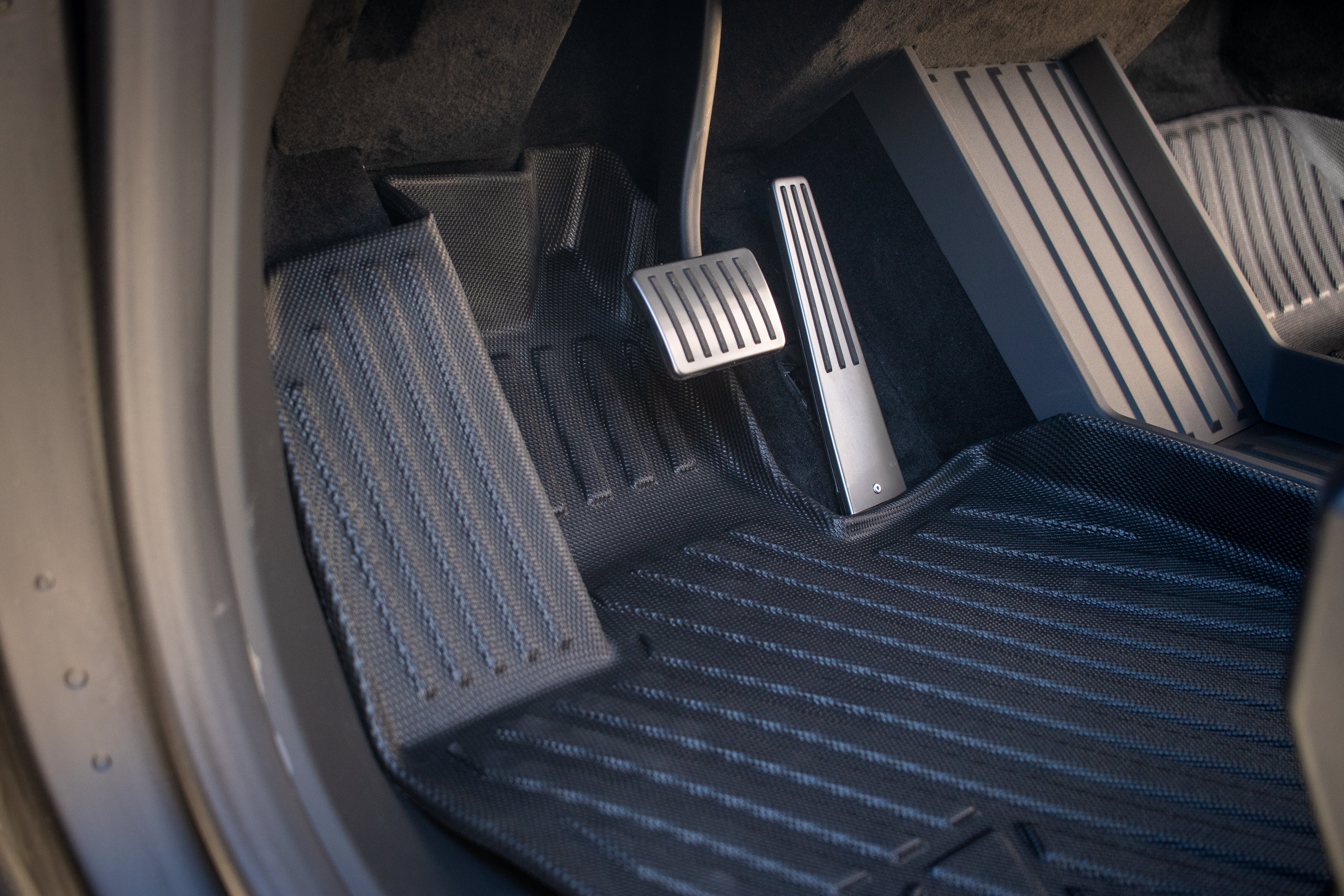 A close-up of the Tesla Cybertruck's driver-side footwell showcases JOWUA All Weather Floor Liners with a textured black design, alongside sleek metal accelerator and brake pedals with a brushed finish and rectangular grip patterns for enhanced control.