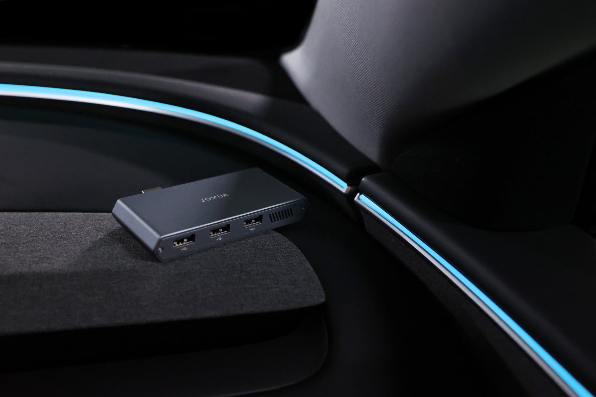 A 3-Port USB DashCam Hub (1TB NVMe SSD) from JOWUA, featuring a black design with a Data Transmission Port, sits on the textured dashboard of a Tesla Model S interior. Blue ambient lighting enhances the car's design, creating a modern and sleek atmosphere.