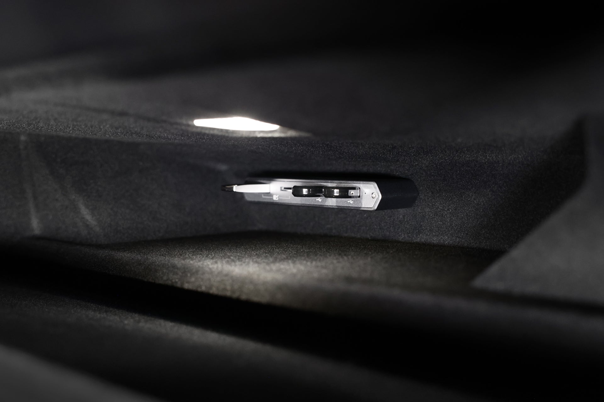 A sleek, modern JOWUA 3-Port USB DashCam Hub (MicroSD Reader) V2 rests in a dimly lit compartment with soft overhead lighting illuminating its metallic surface, designed for Tesla owners' data port compatibility. The surrounding interior is dark and plush, highlighting the hub's elegant design.