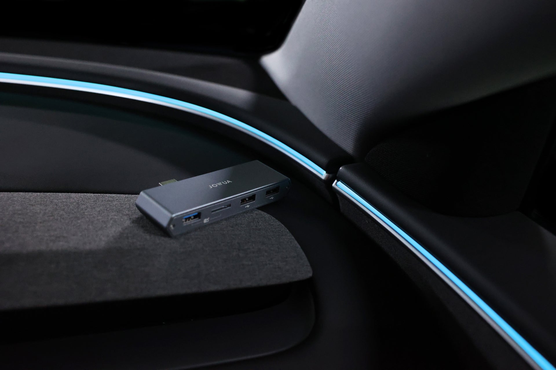 Positioned on a textured surface inside a car, the JOWUA Dual-Function Storage HUB (MicroSD Reader & Tesla USB) provides data port expansion with multiple ports, such as USB-A, HDMI, and USB-C. The interior highlights ambient blue lighting along the edges, making it an ideal Tesla accessory for tech-savvy drivers.