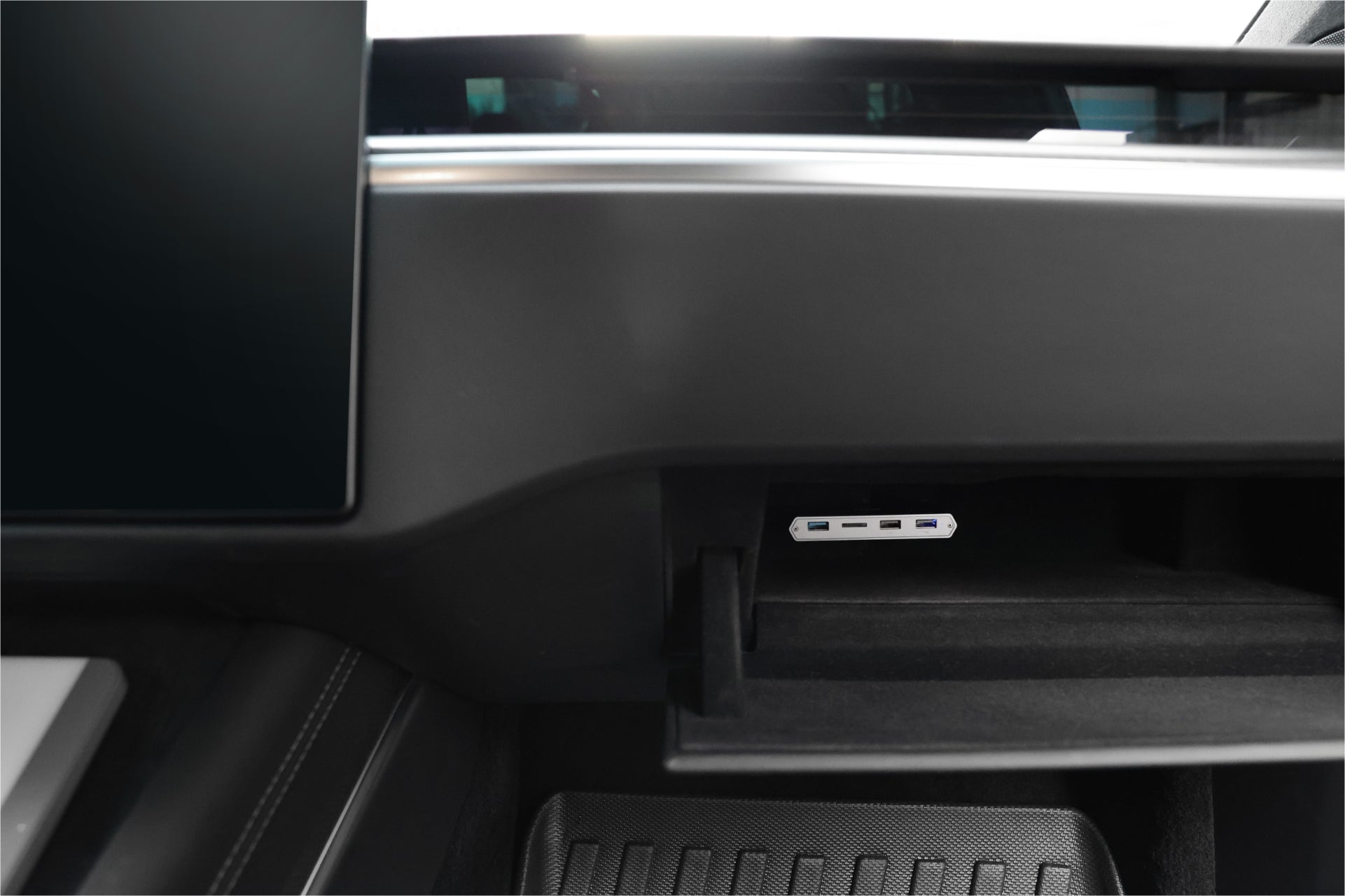 A close-up of a car's interior reveals an open storage compartment under the dashboard, featuring the JOWUA 3-Port USB DashCam Hub (MicroSD Reader) V2, perfect for Tesla owners needing data port connections. The dashboard is sleek and modern, with a large screen visible on the left.