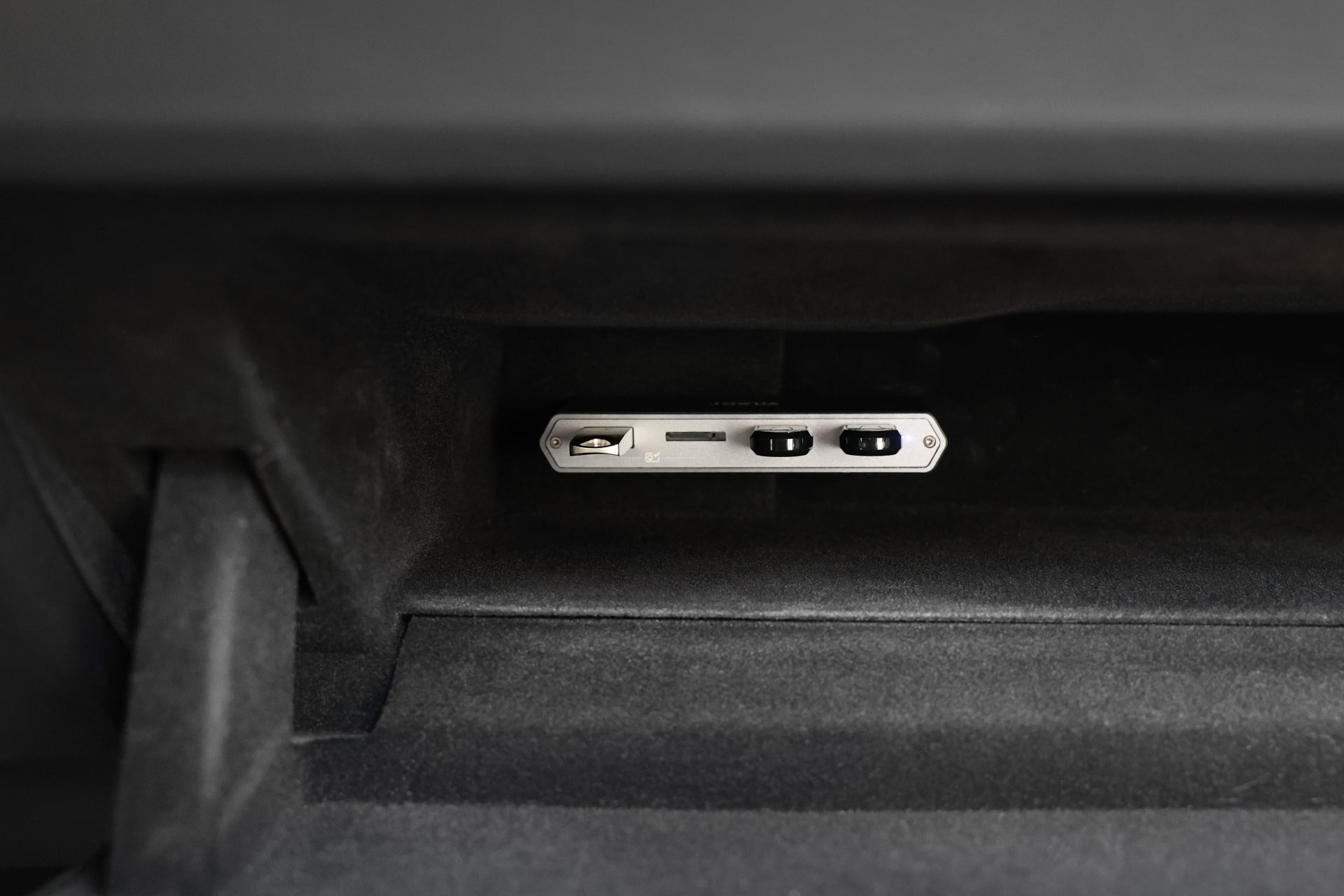 A close-up view of a car's interior glove compartment reveals a JOWUA accessory: the Dual-Function Storage HUB, which includes an illuminated wireless charging dock featuring two Tesla USB ports and a MicroSD reader, designed to prevent electromagnetic interference, all set against a dark fabric background.