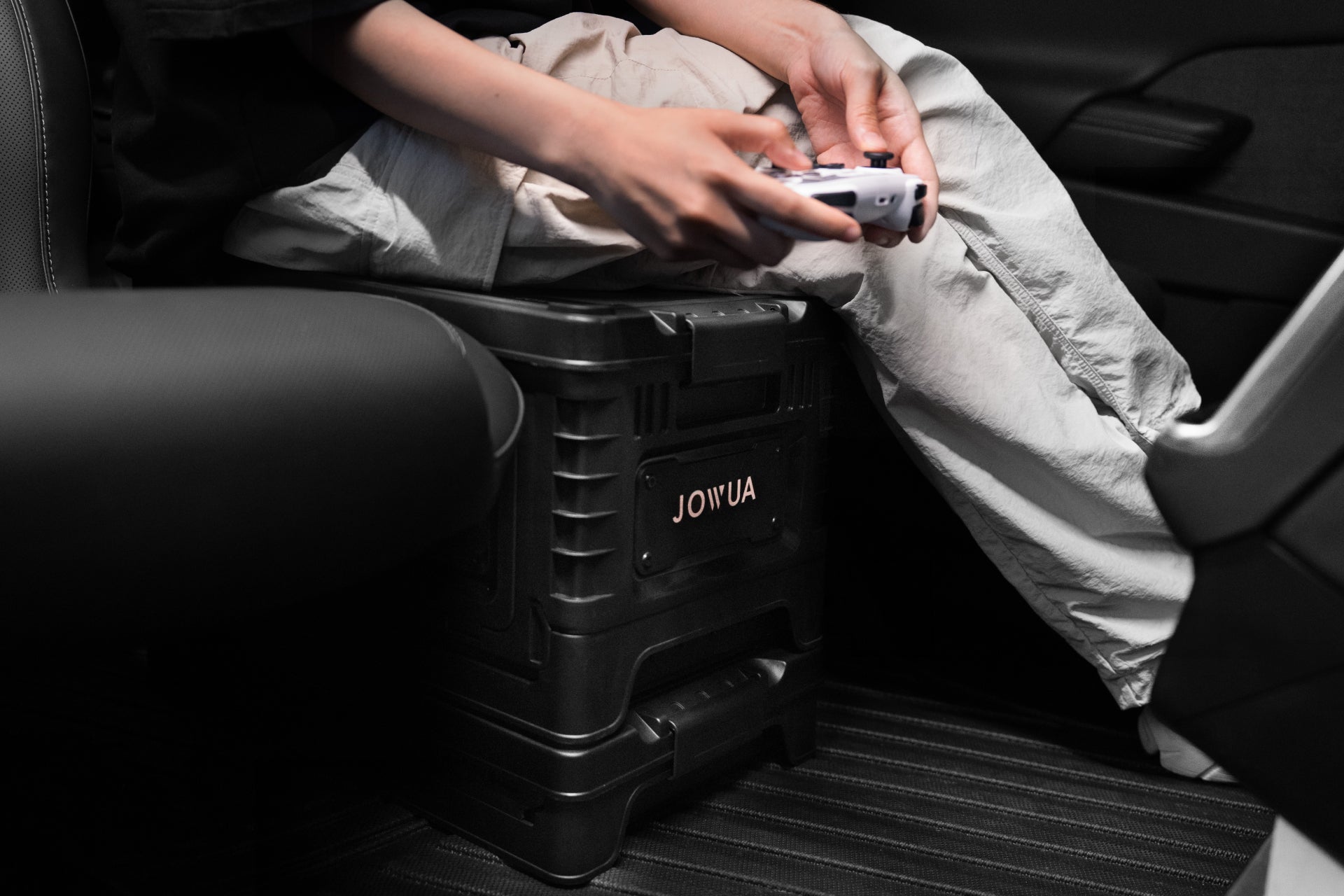 Inside the vehicle, a person holds a gaming controller, their hands resting on a sturdy 30L Stackable Storage Box and Tesla Trunk Organizer with the "JOWUA" brand. They're wearing light-colored pants and a dark top.