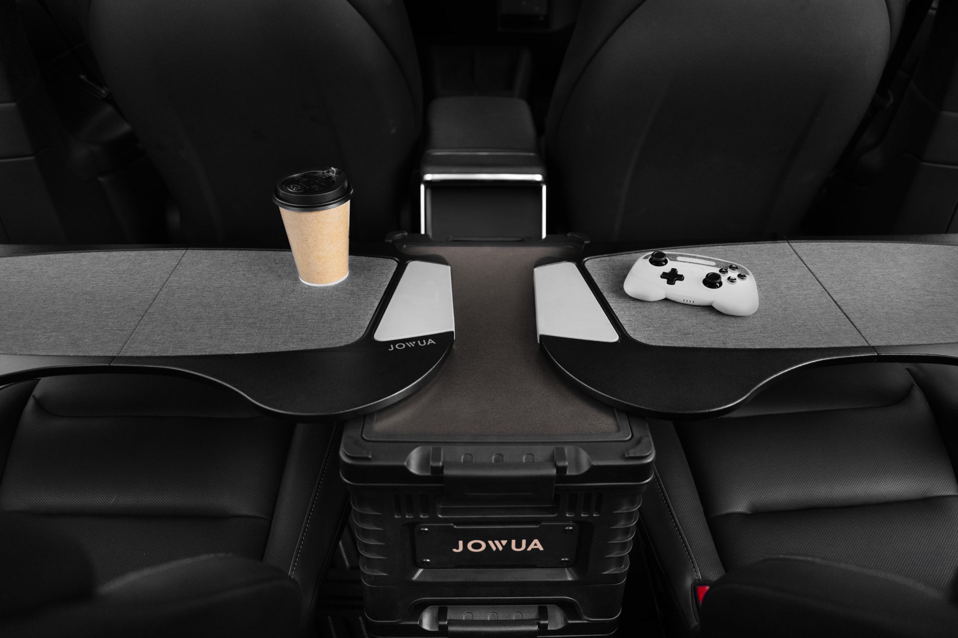 A car interior showcases a console setup with a coffee cup on the left and a gaming controller on the right. The JOWUA 30L Stackable Storage Box and Trunk Organizer for Teslas enhances the customizable tray tables, creating a personalized space for activities while traveling.