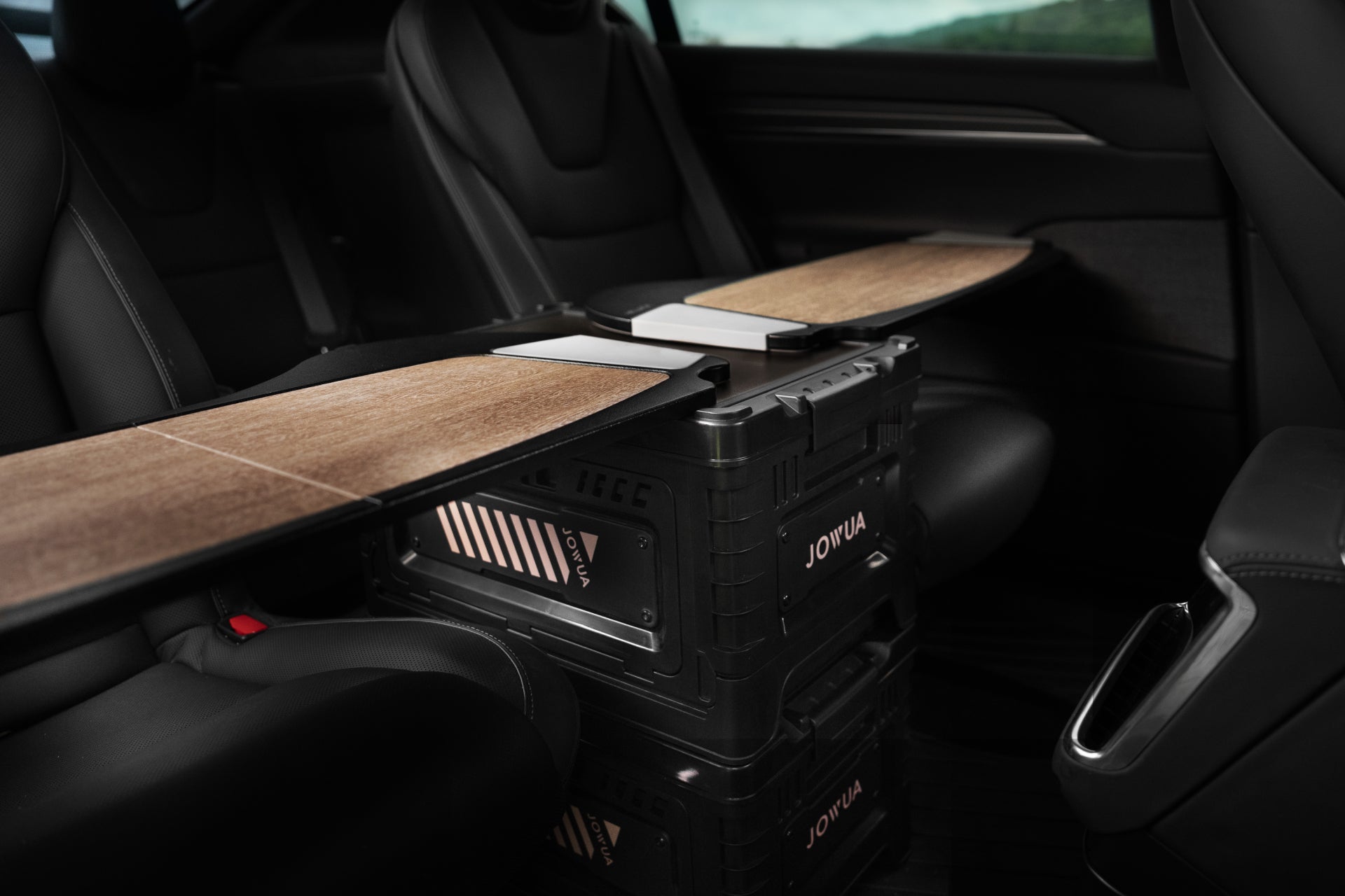 The interior of a car showcases a snowboard placed on top of two stacked, black JOWUA brand storage boxes, which boast a robust stackable design. The vehicle might be equipped with the 30L Stackable Storage Box and Trunk Organizer for Teslas, featuring black leather seats with minimal natural light streaming through the window.