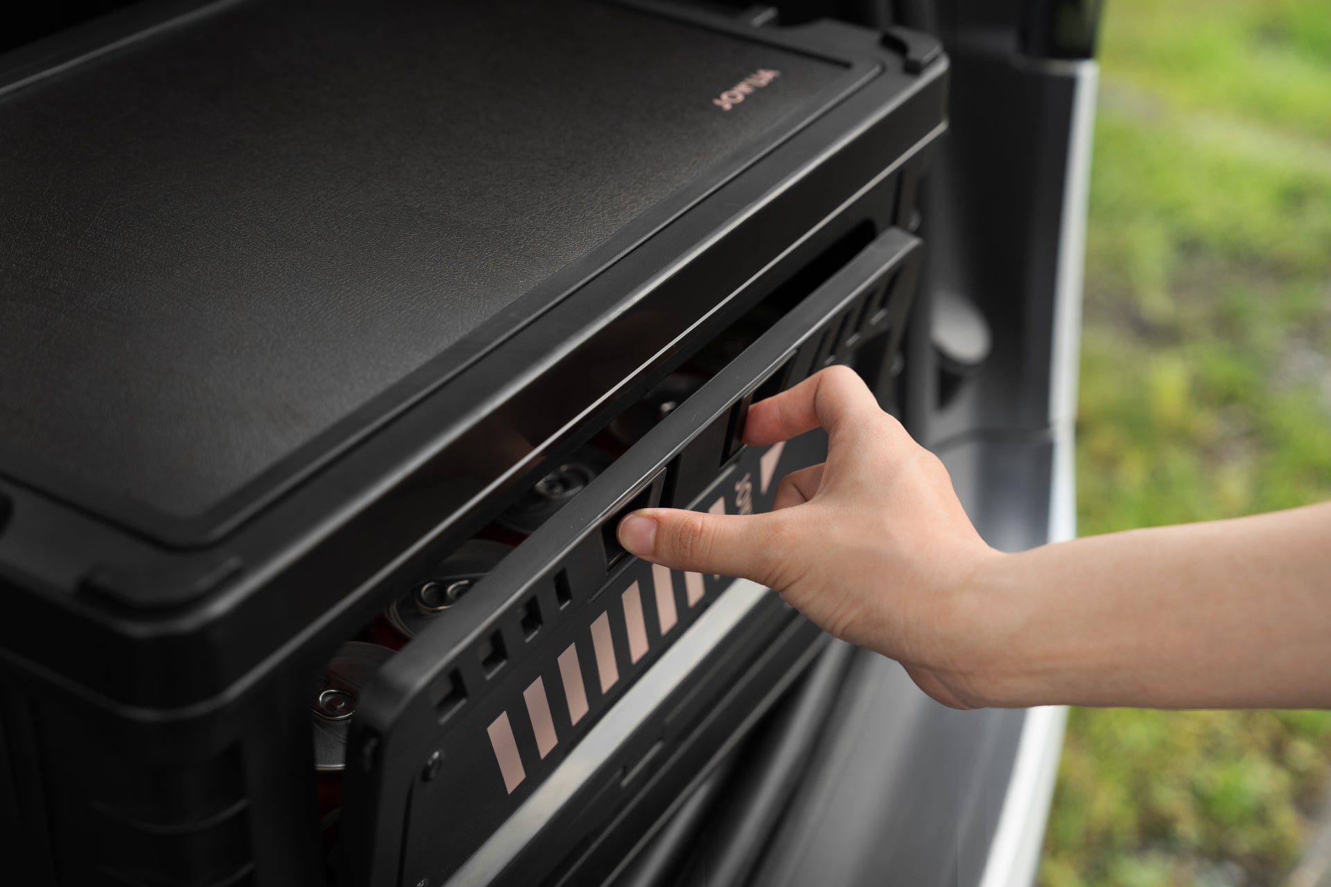 A person's hand opens a JOWUA 30L Stackable Storage Box and Trunk Organizer for Teslas in the back of a vehicle. The organizer is outdoors, with green grass visible in the background.