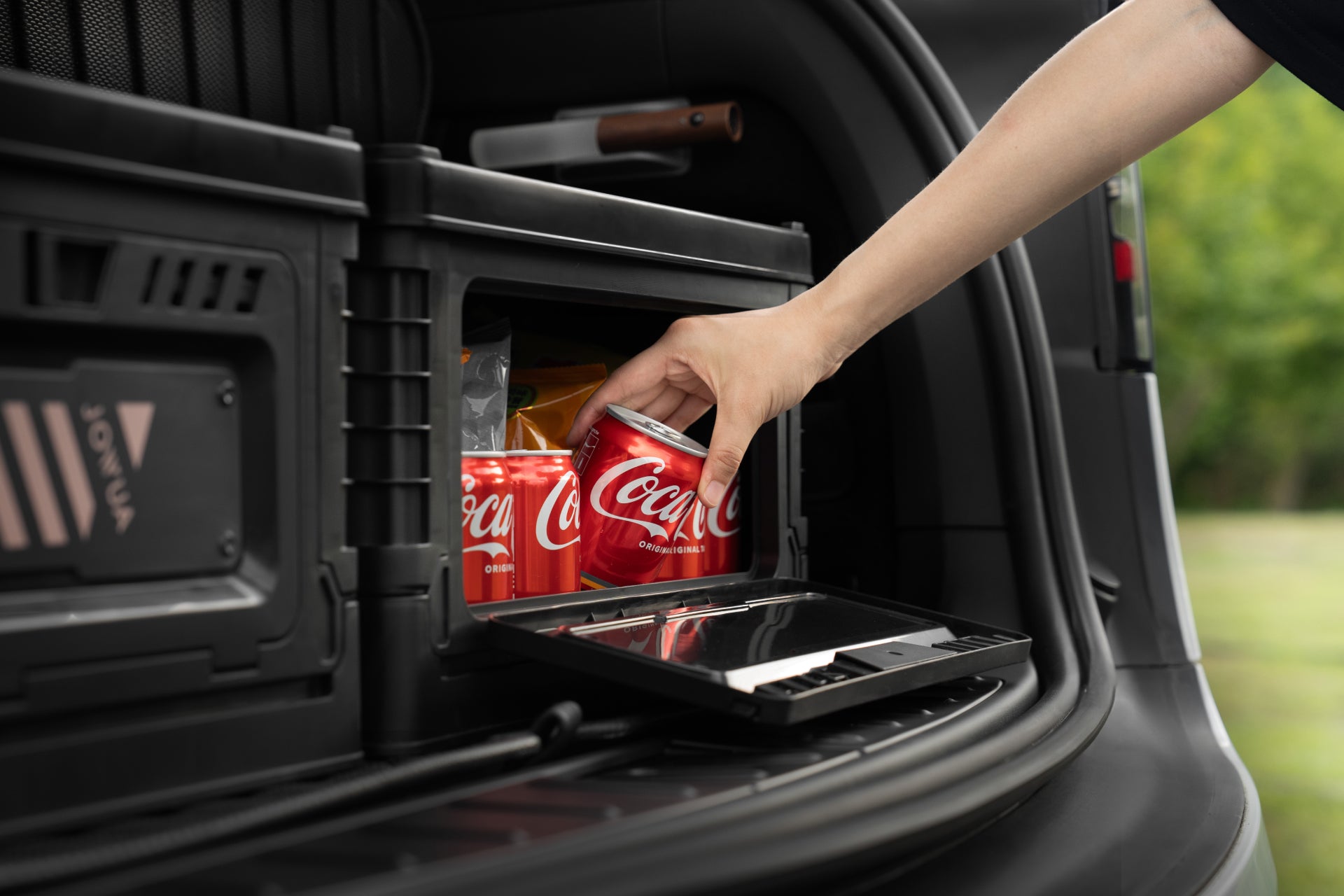 A hand reaches into a JOWUA 30L Stackable Storage Box and Trunk Organizer for Teslas to grab a red can of Coca-Cola among several others, with blurred greenery in the background hinting at an outdoor setting.