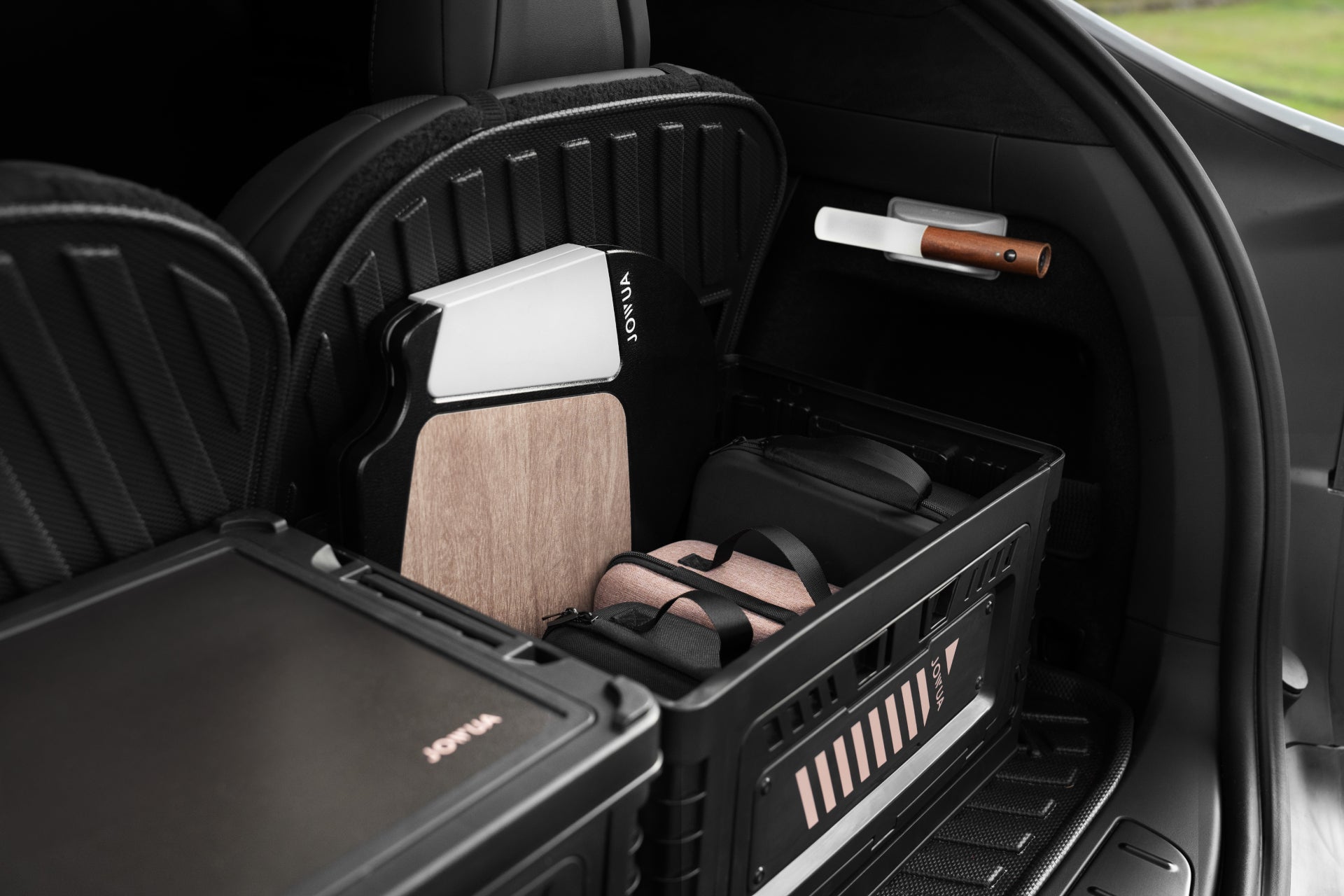 A JOWUA 30L Stackable Storage Box and Trunk Organizer enhances a Tesla's sleek interior by neatly arranging various items, including a brown and beige cutting board, a pink bag, and a white rolled-up mat. Its heavy-duty, stackable design maximizes space utilization, with folded seats offering additional room.