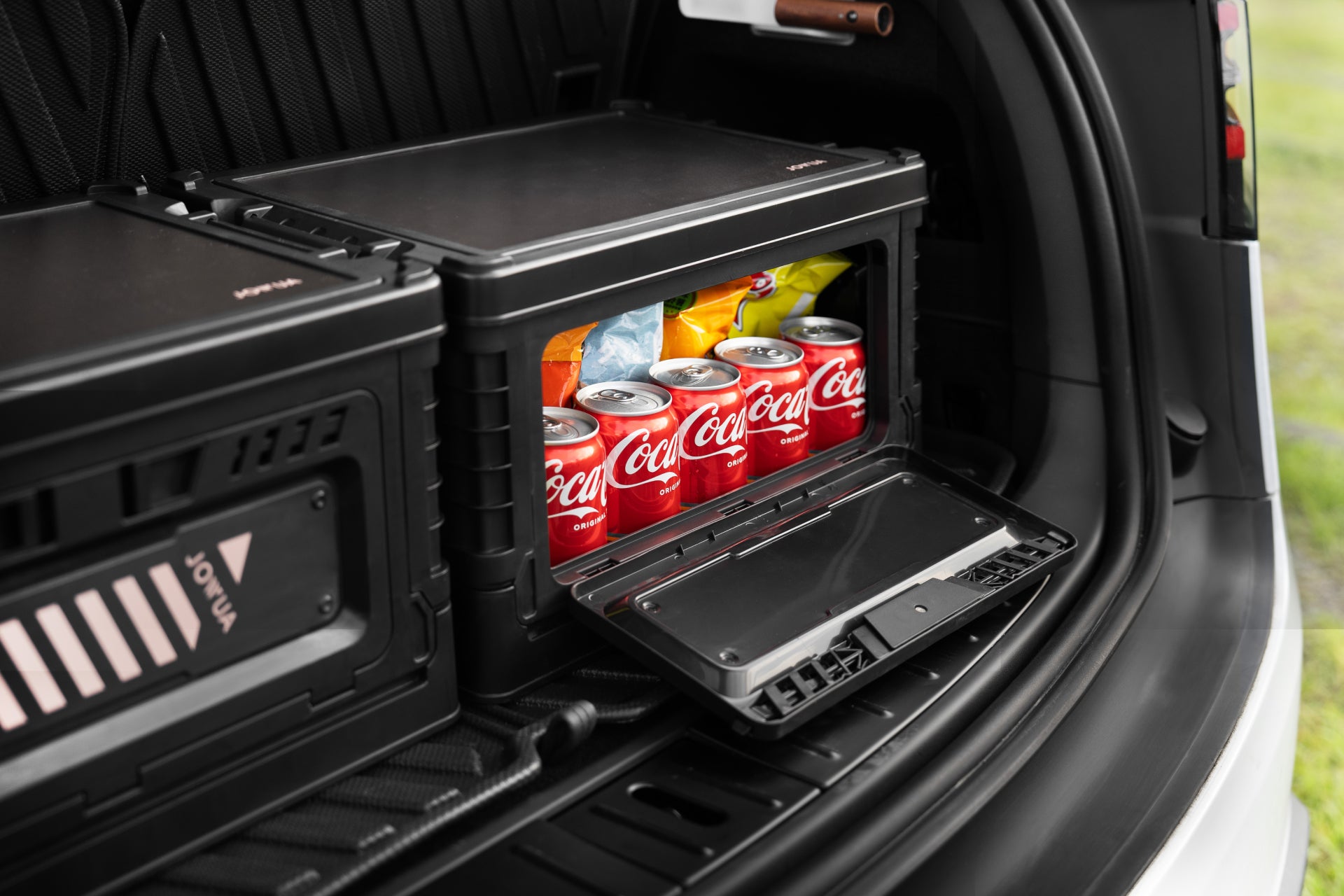 An open JOWUA 30L Stackable Storage Box and Trunk Organizer for Teslas in a car showcases several red Coca-Cola cans neatly aligned beside vibrant packaged snacks. A closed compartment is nearby, creating an ideal setup for a road trip.