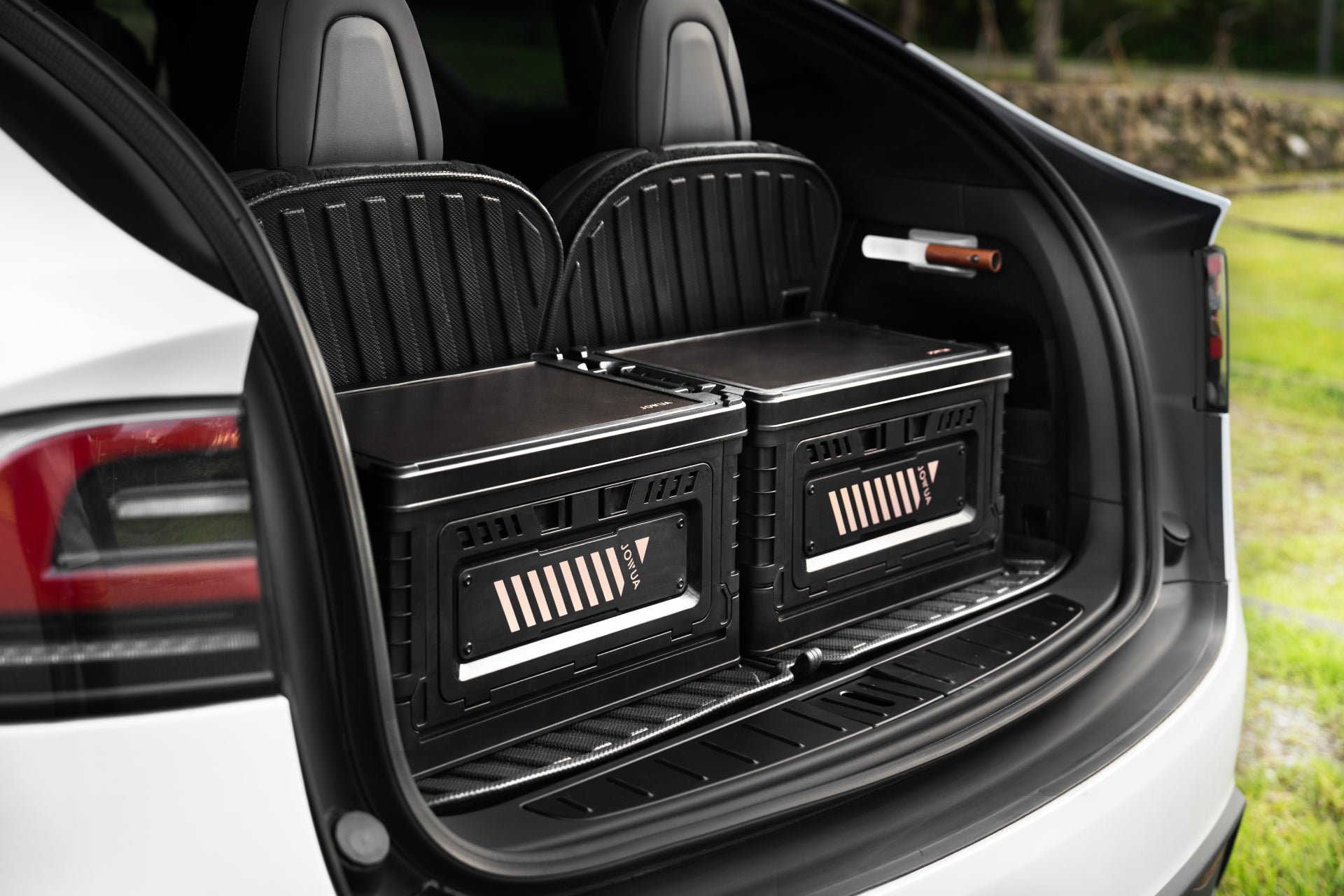 A white electric car's trunk houses two large black battery packs, neatly arranged next to a JOWUA 30L Stackable Storage Box and Trunk Organizer for Teslas. The rear seats can be seen, complemented by the serene setting of a green field in the background.