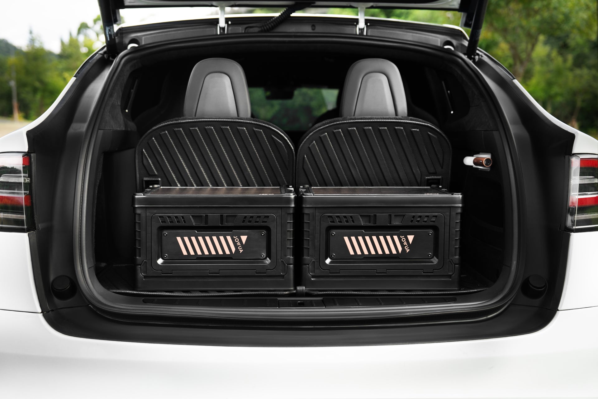 The image features the open trunk of a white car, showcasing two substantial JOWUA 30L Stackable Storage Boxes and Trunk Organizers, designed for Teslas. The back seats are visible, with trees in the background.