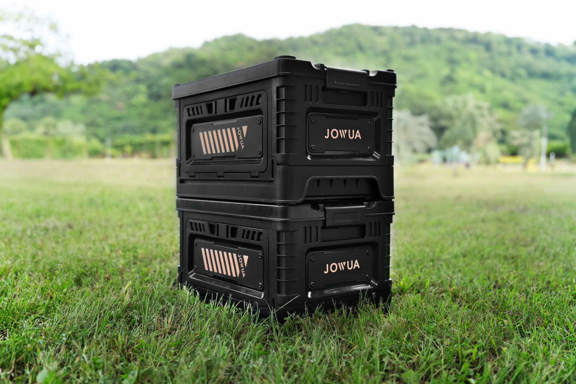 Two JOWUA black stacked storage boxes and trunk organizers, designed specifically for Teslas, sit on a grassy field with trees and hills in the background. Their sturdy, compact design makes them ideal for outdoor adventures.