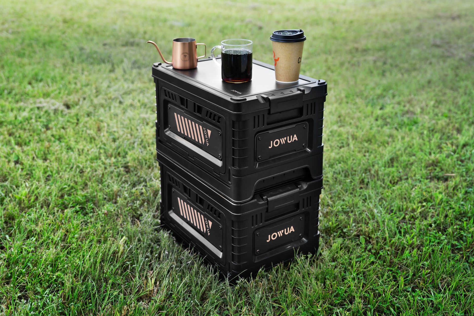 Two JOWUA 30L Stackable Storage Boxes, perfect for organizing a Tesla trunk, are positioned on the grass to create an impromptu table. A metal kettle, a glass mug filled with black coffee, and a paper coffee cup with a lid sit on top.