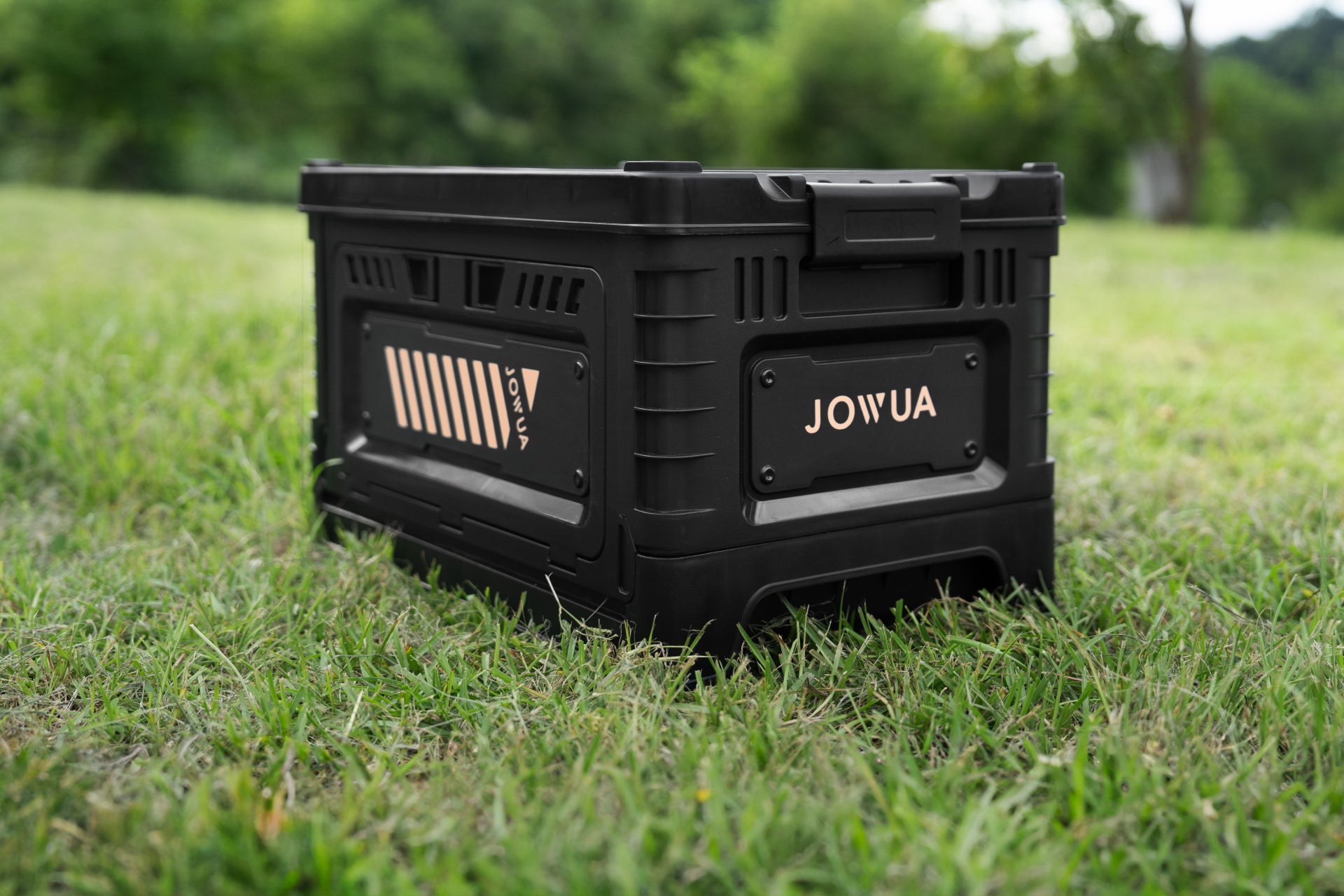 A 30L Stackable Storage Box and Trunk Organizer for Teslas, branded by JOWUA, sits on green grass outdoors, mirroring the style of a Tesla trunk organizer. Trees and blurred greenery in the background create a natural setting. Equipped with sturdy handles and reinforced edges, this storage box is perfect for rear seat storage or use as a car interior organizer.